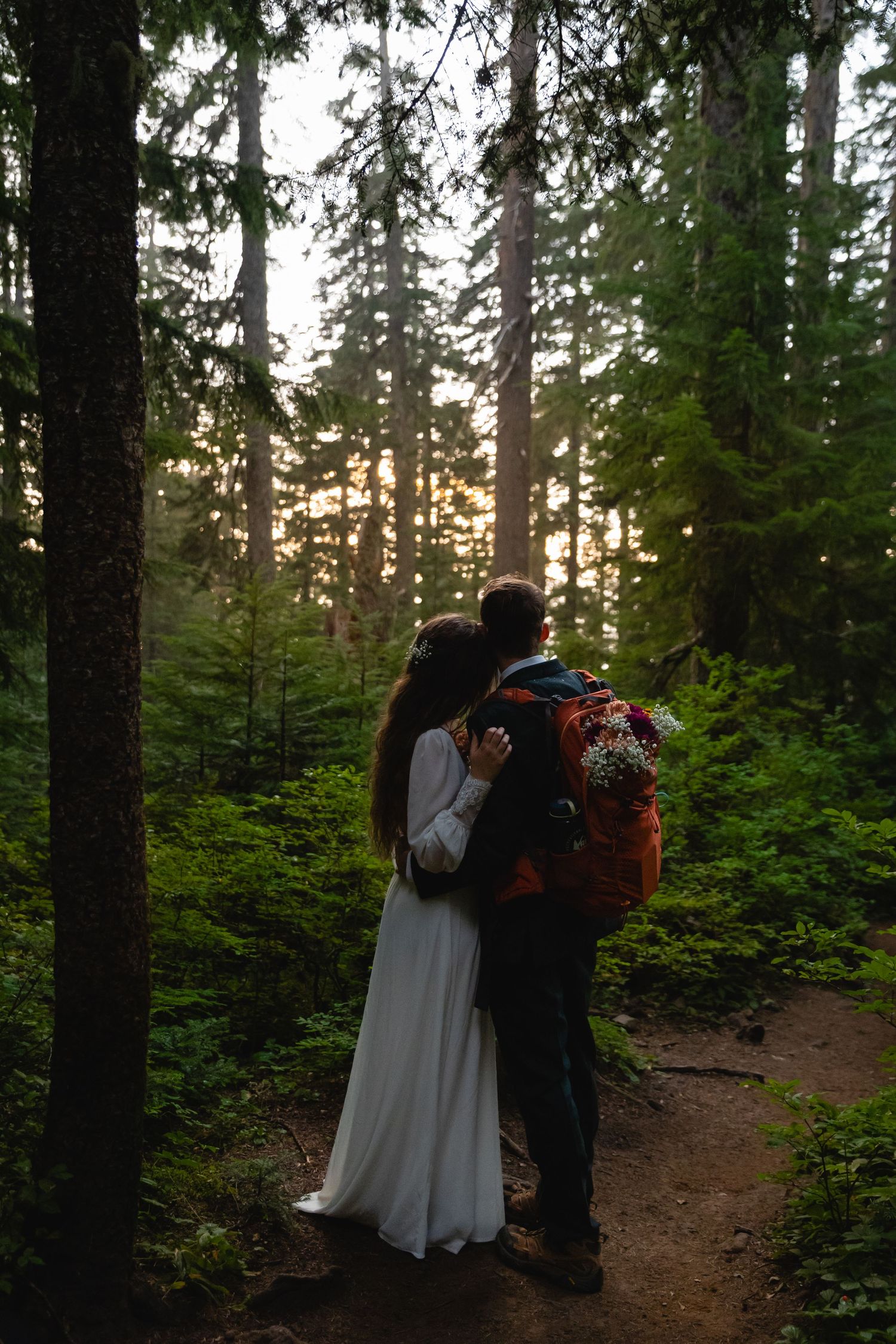 A couple in hiking attire stands on a forest trail at sunset, surrounded by evergreen trees.