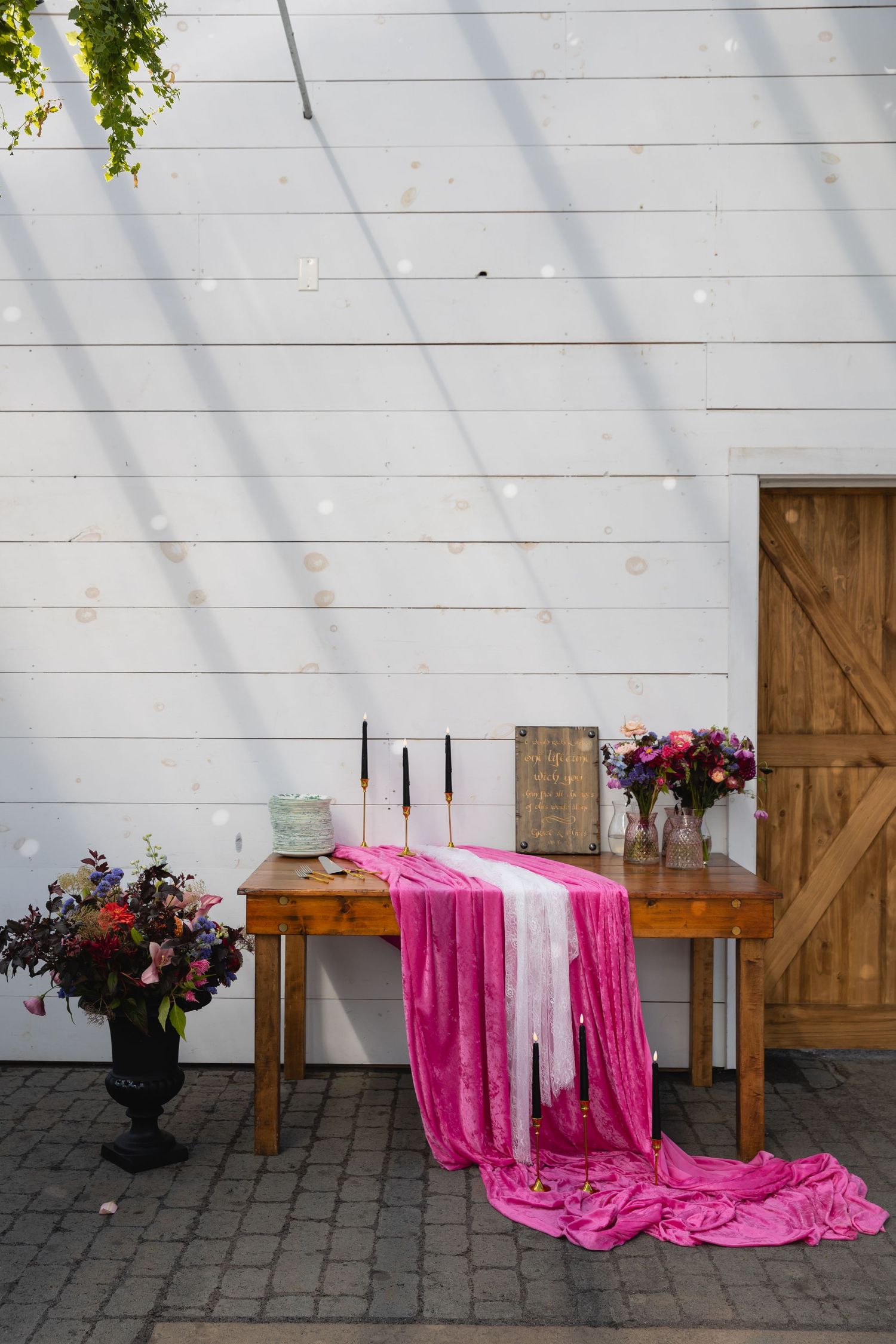 Rustic wooden table with flowing pink fabric and floral arrangements against white shiplap wall and barn door.