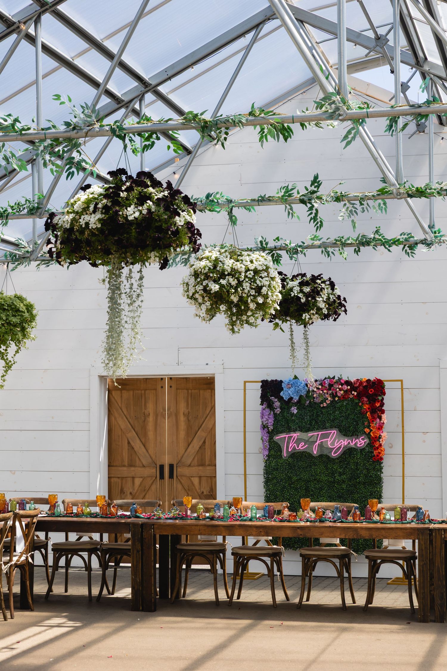 Long wooden farm tables set up in a greenhouse venue with hanging floral arrangements and rustic barn doors.
