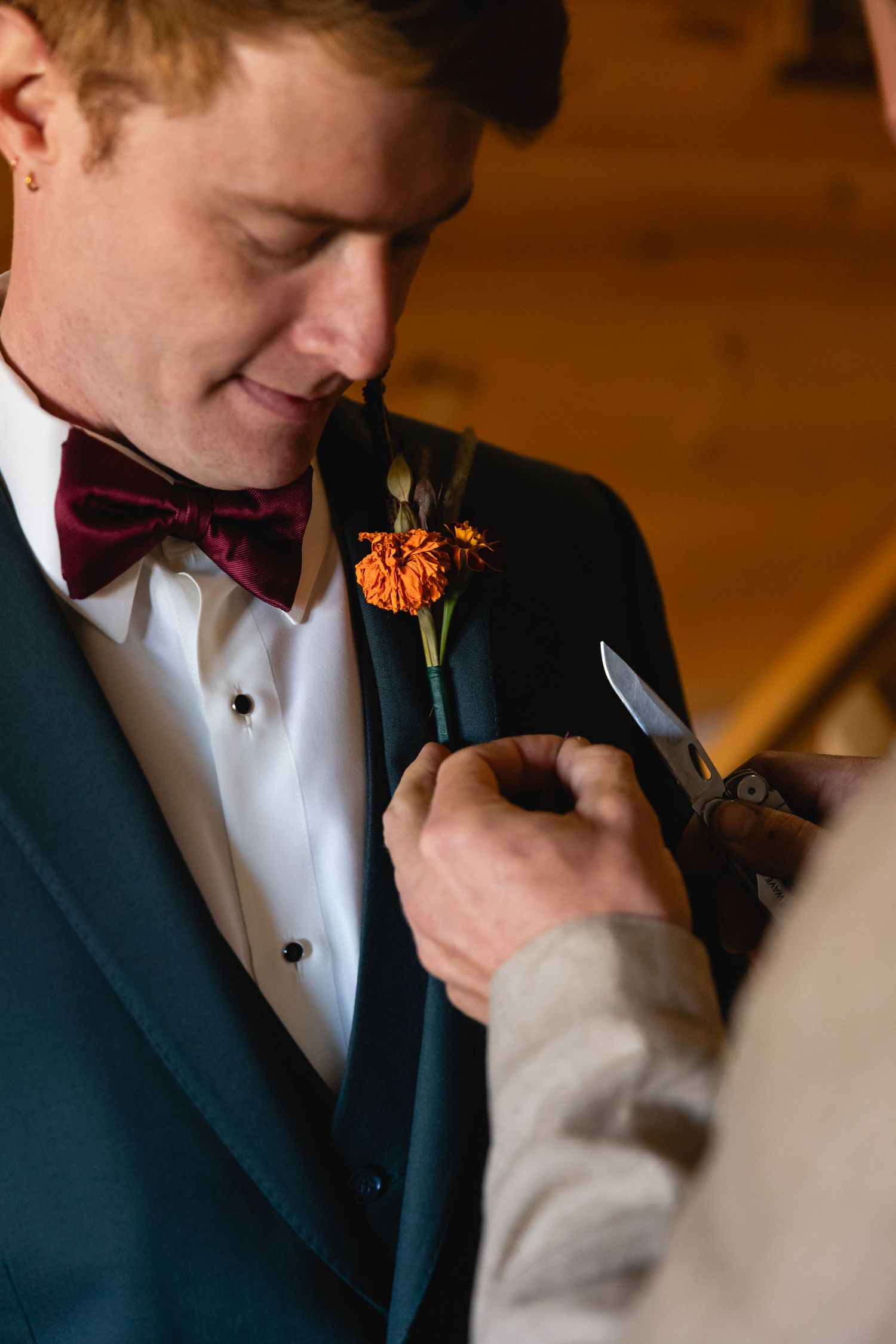 Someone adjusts an orange boutonniere on a groom wearing a burgundy bow tie and dark teal suit jacket.