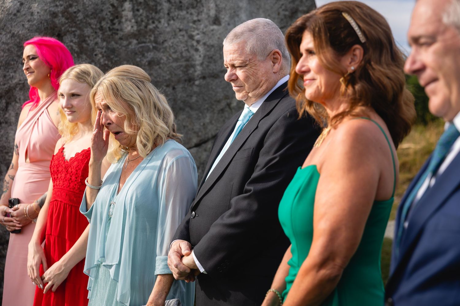 Wedding ceremony attendees stand in formal attire while watching an outdoor celebration.