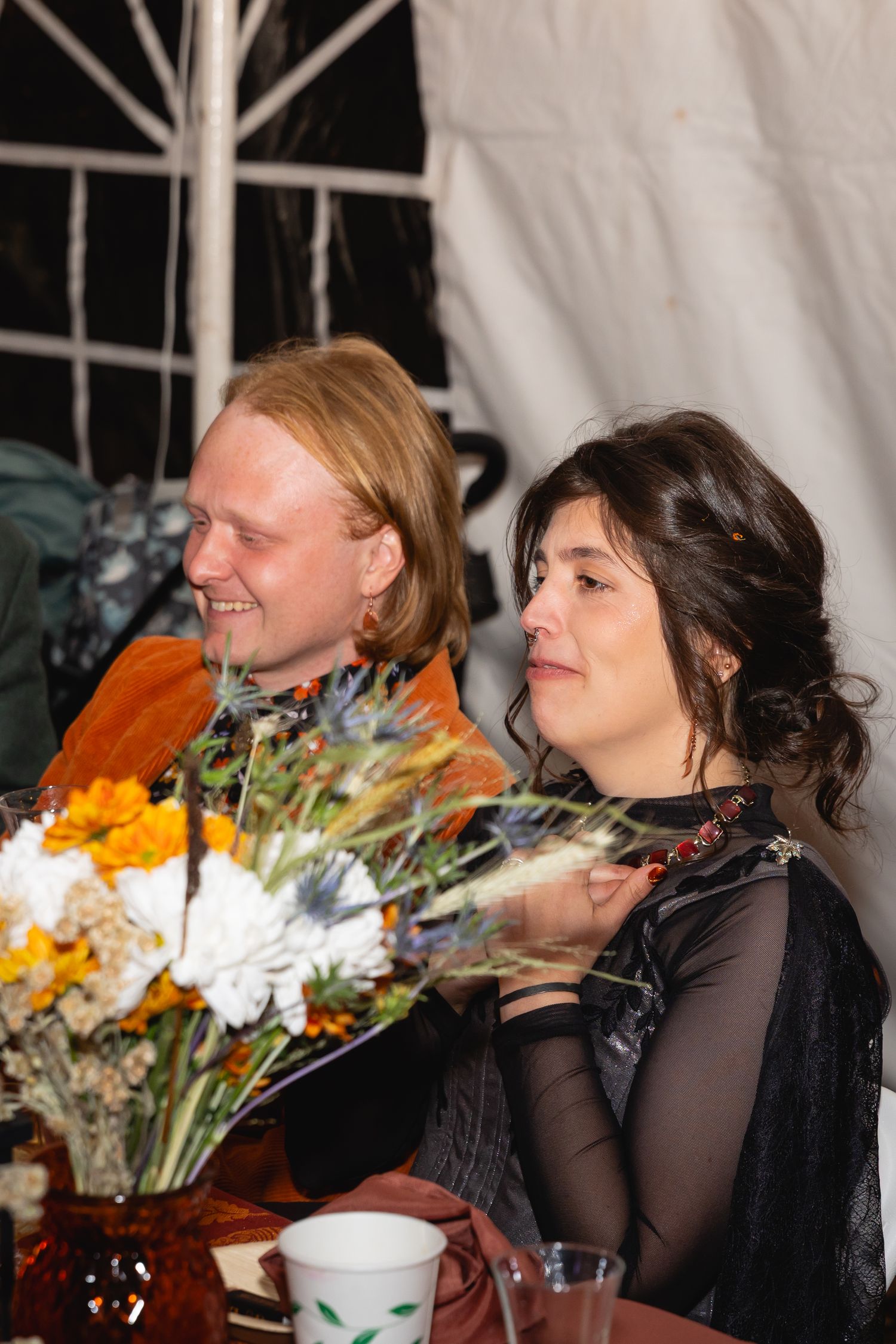 Two people share a laugh at a dinner table with colorful flower arrangements in the background.