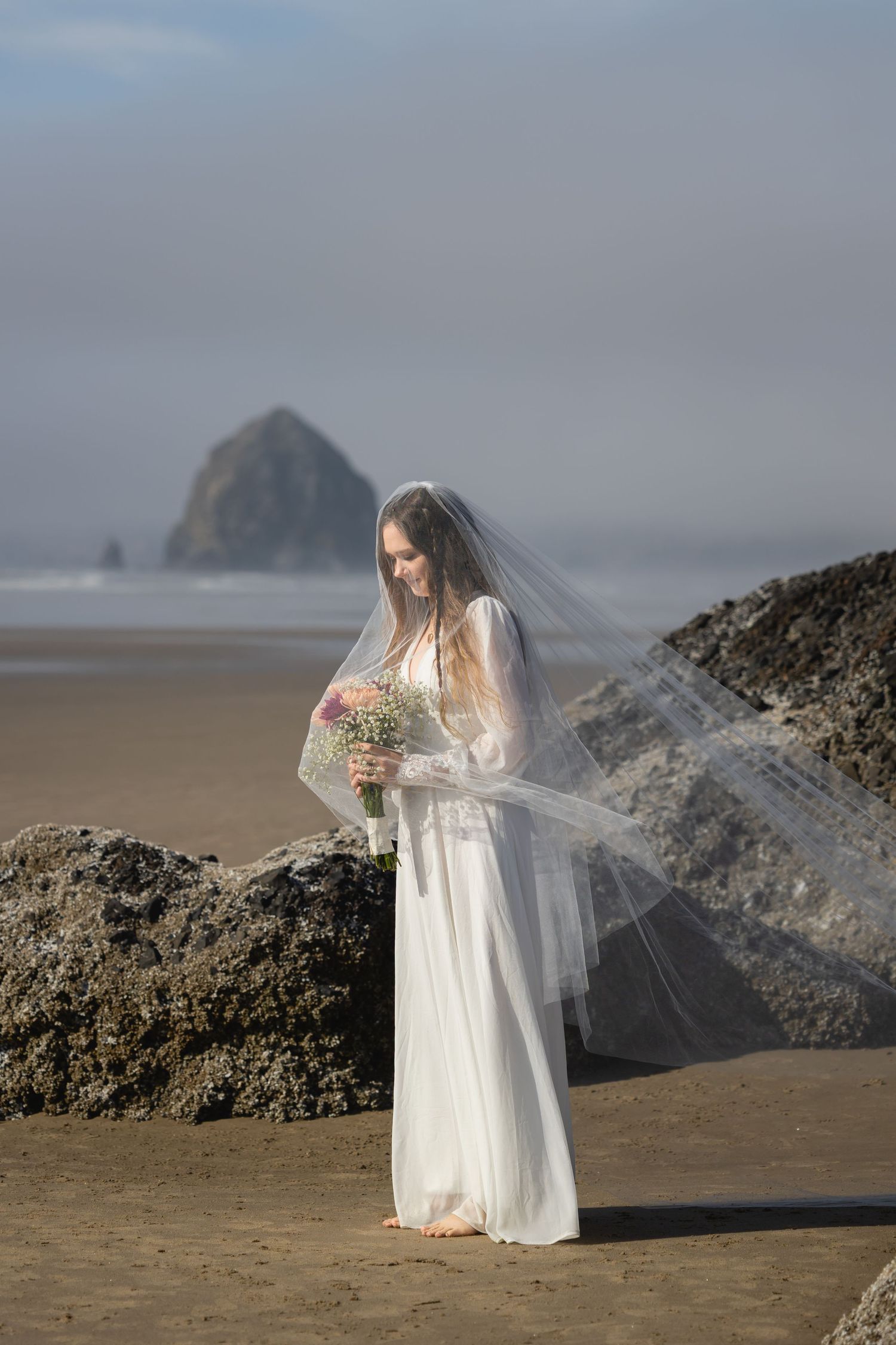 A bride in a flowing white dress and veil holds a bouquet on a misty beach with Haystack Rock visible in the background.