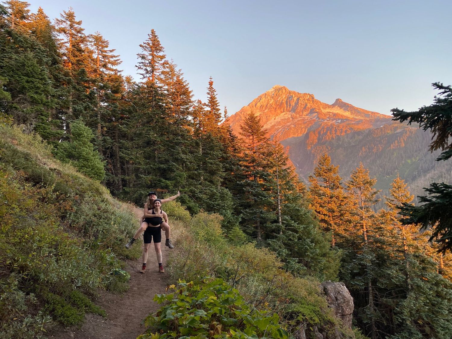 Hiker with backpack on mountain trail at golden sunset with illuminated peak through pine trees.