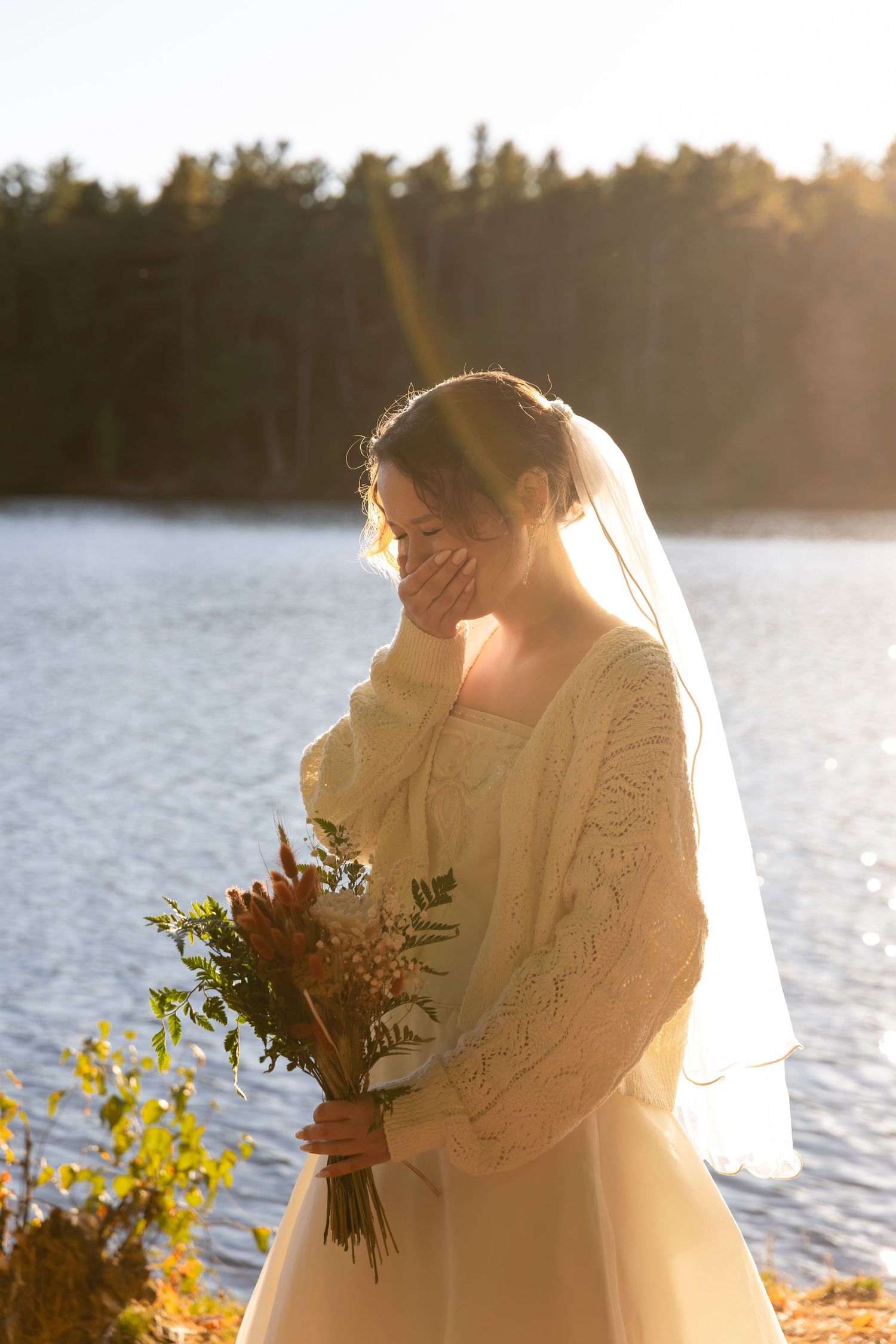 Emotional bride in a lace dress and veil holds a bouquet while standing by a sunlit lake at golden hour.