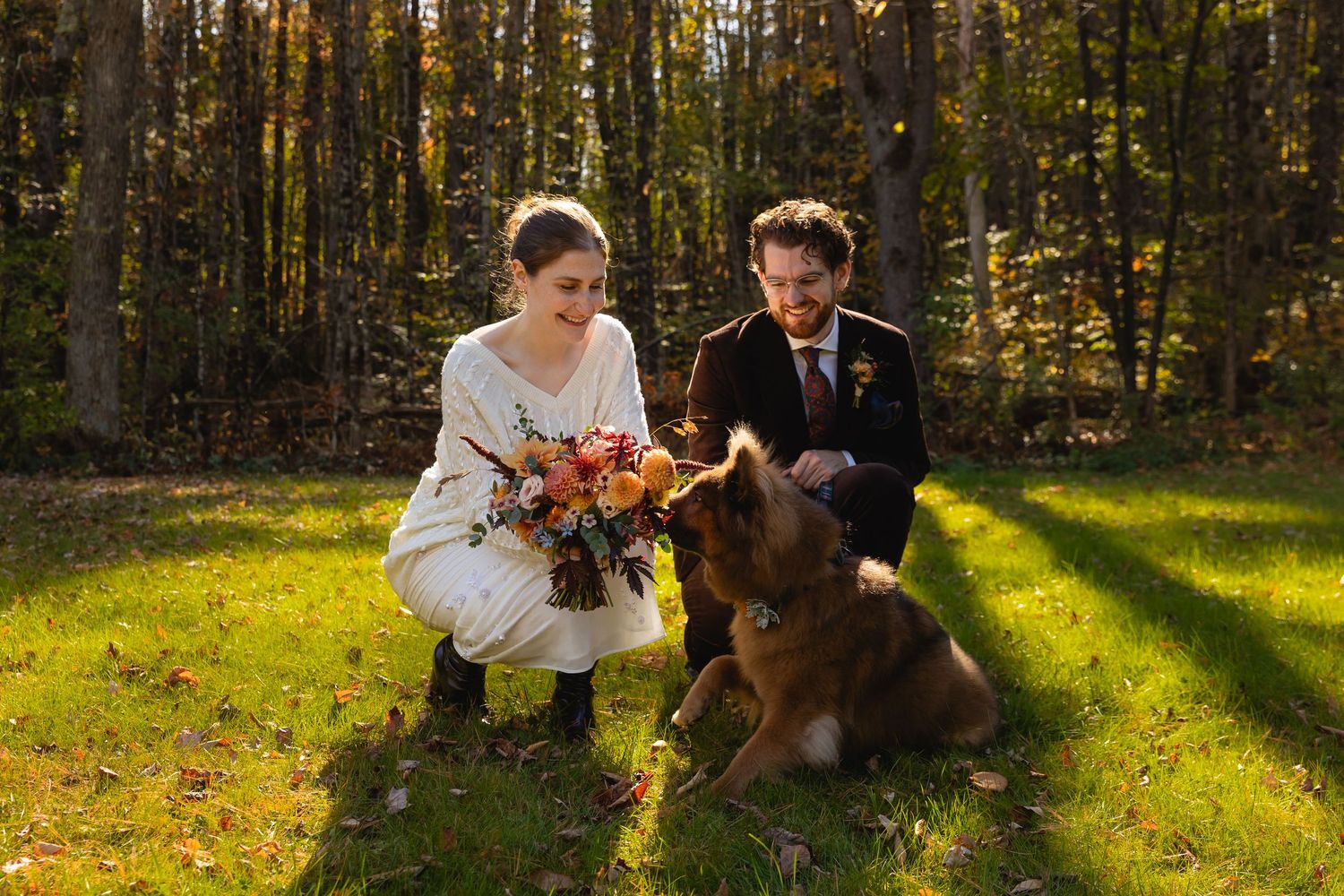 Bride and groom kneel in sunlit forest clearing, petting their brown dog while holding a fall bouquet.