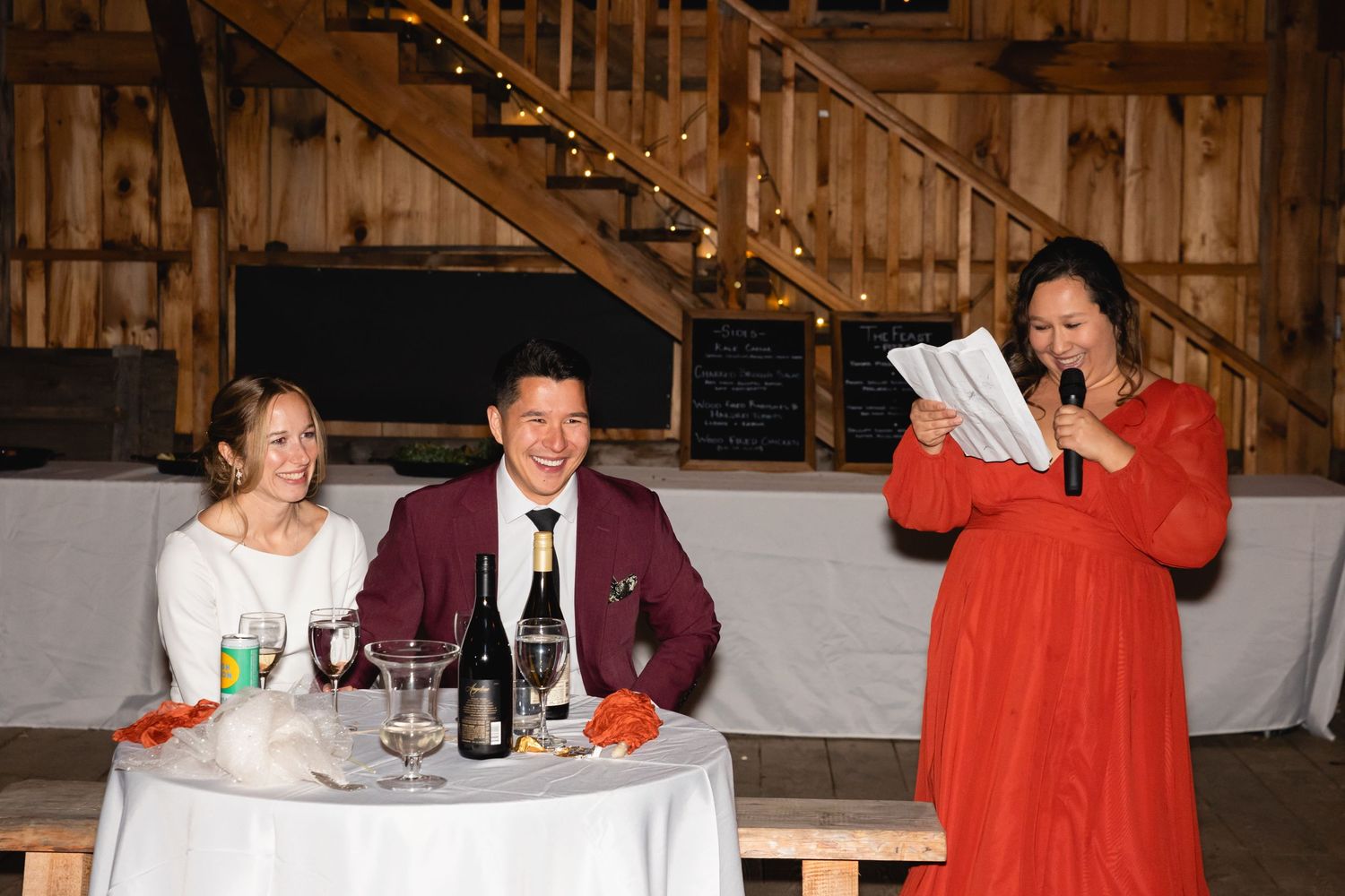 People gather at a rustic barn wedding reception with wooden decor and string lights overhead.