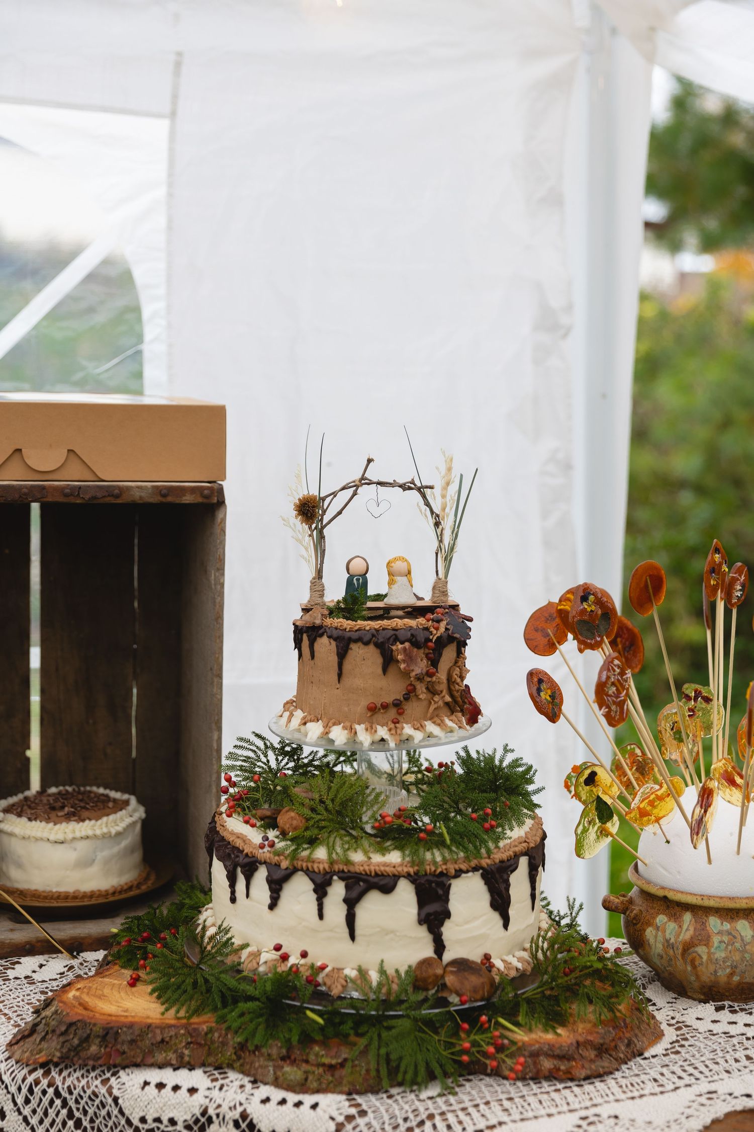 Rustic two-tier wedding cake decorated with twigs, greenery and chocolate drip on a wood slice.