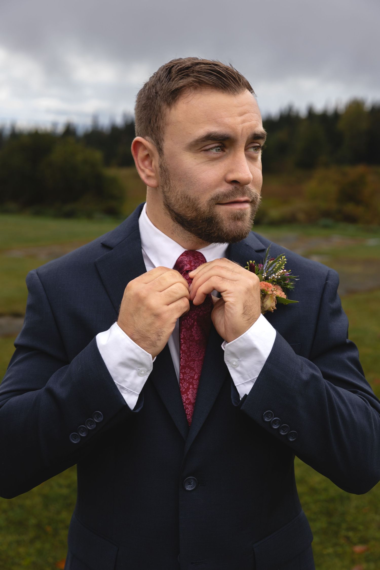 A groom adjusts his burgundy tie while wearing a navy suit outdoors against a mountainous backdrop.