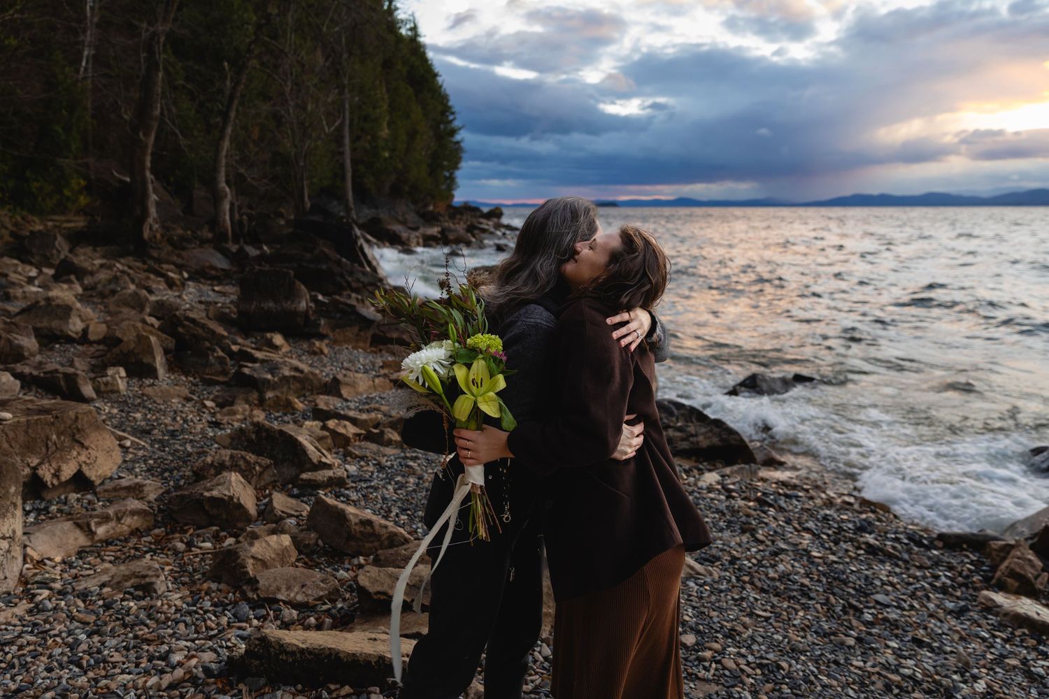Two people share a romantic embrace on a rocky coastline at sunset with dramatic storm clouds overhead and evergreen trees nearby.