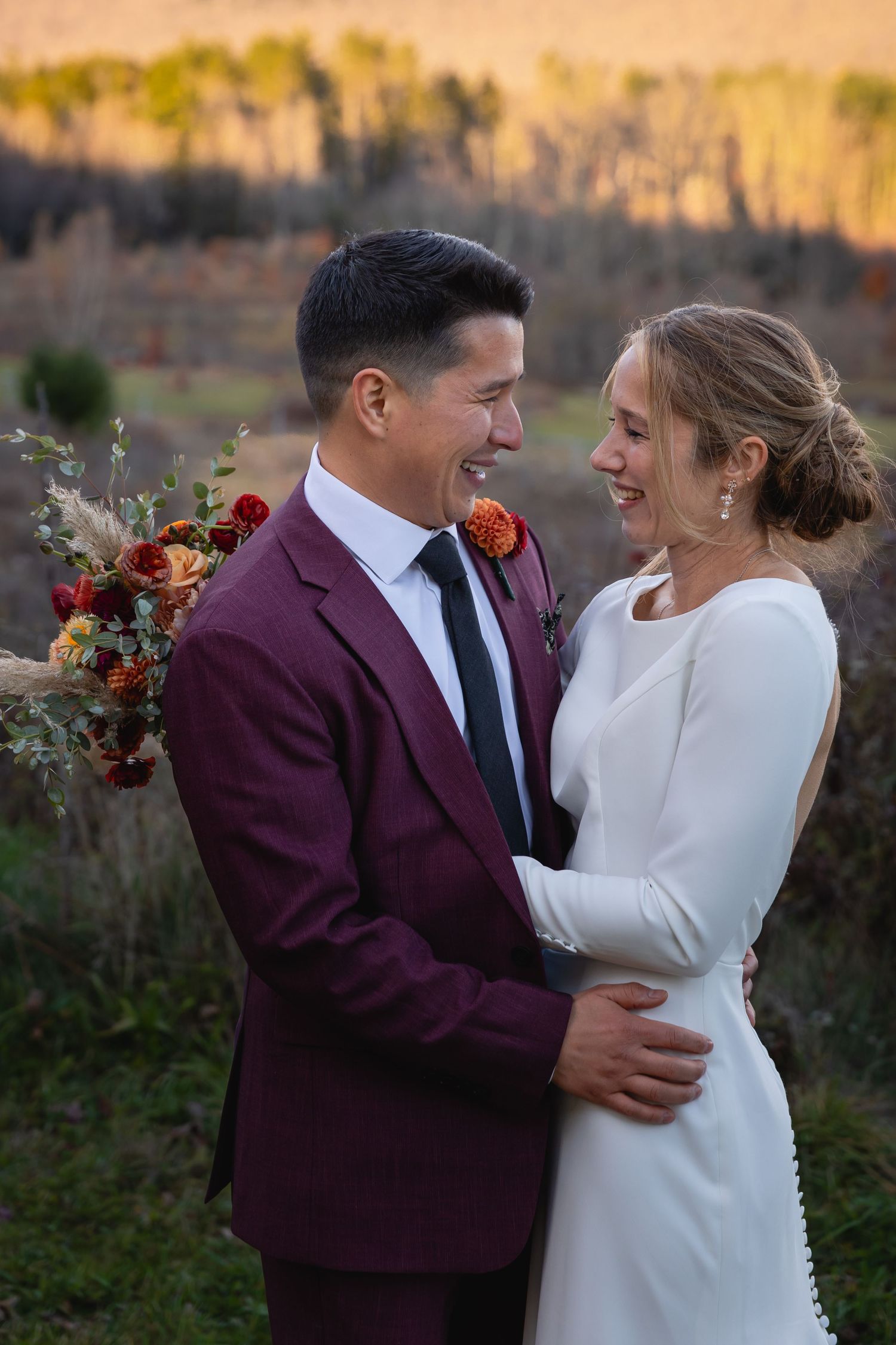 A couple in wedding attire embrace outdoors at sunset, with burgundy suit and white dress against an autumn backdrop.