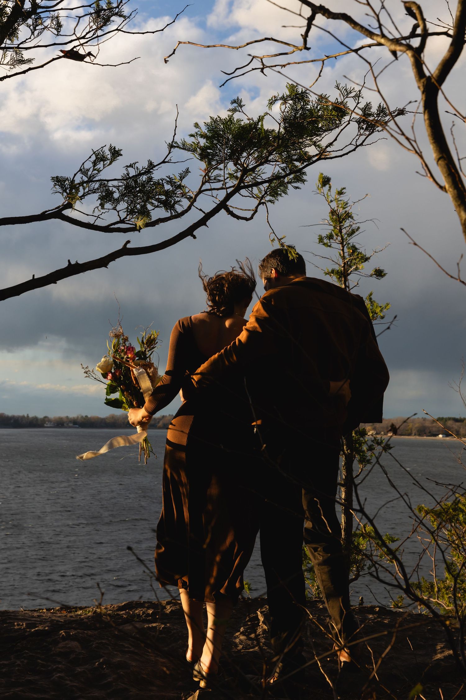 A couple stands silhouetted against a waterfront at sunset, surrounded by tree branches and golden light.