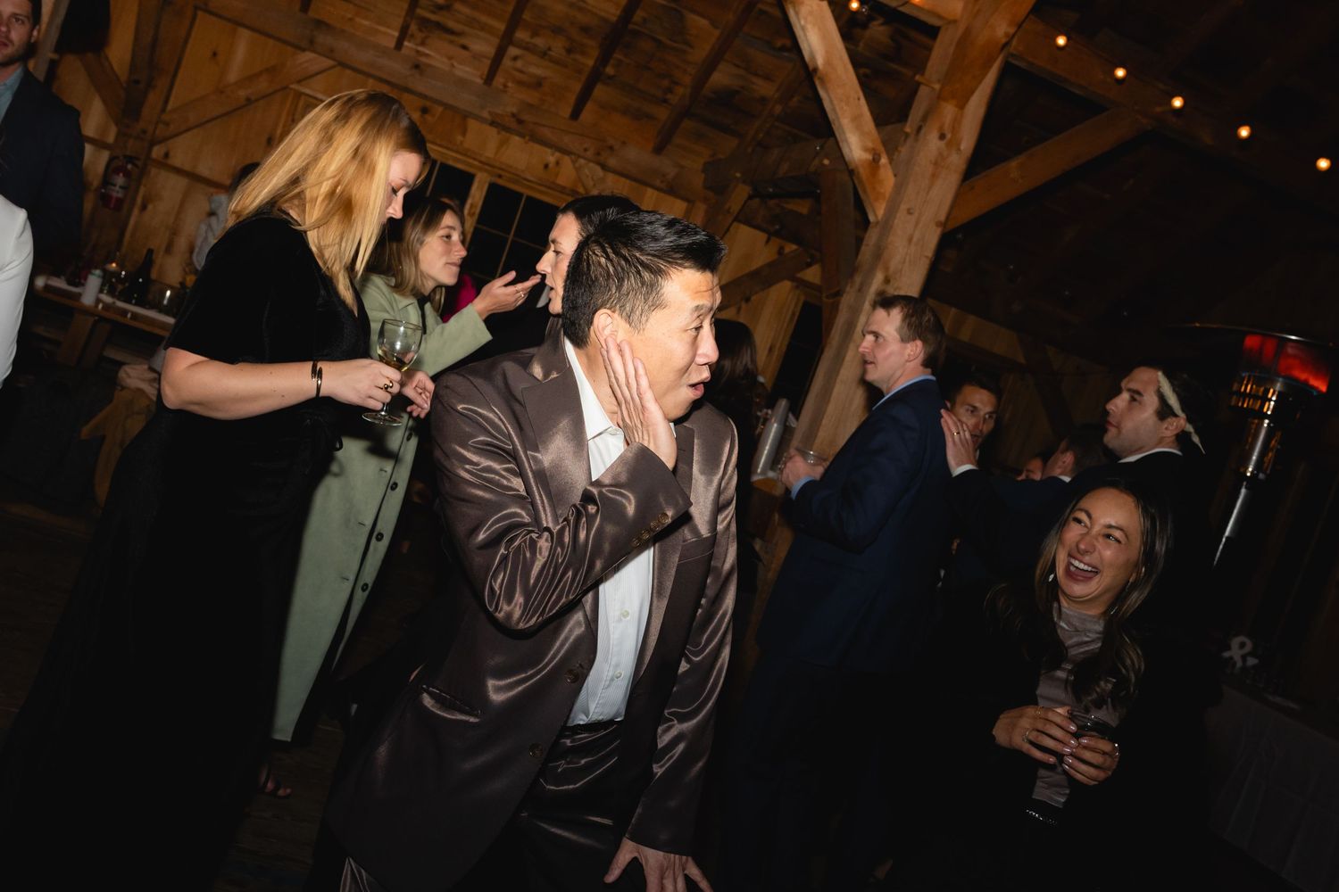 Party guests dance and celebrate together inside a rustic wooden barn with string lights overhead at night.