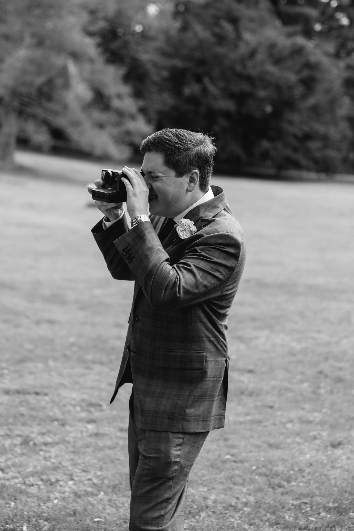 A photographer in a plaid suit jacket takes photos outdoors on a grassy field in black and white.