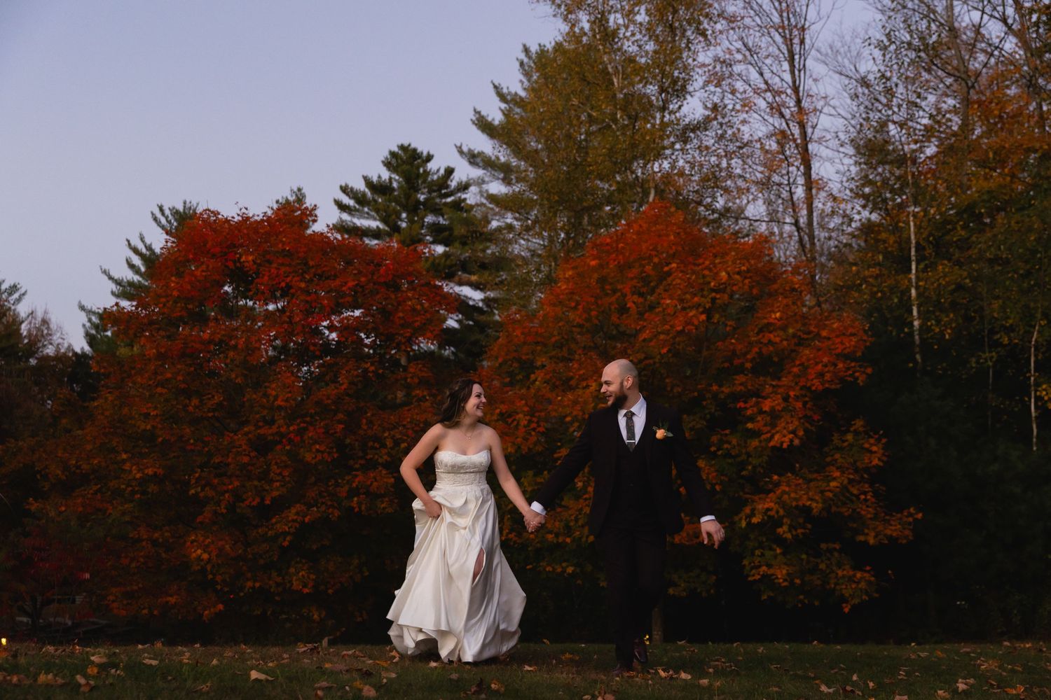 A couple in wedding attire runs joyfully through autumn foliage with vibrant red maple trees in the background.