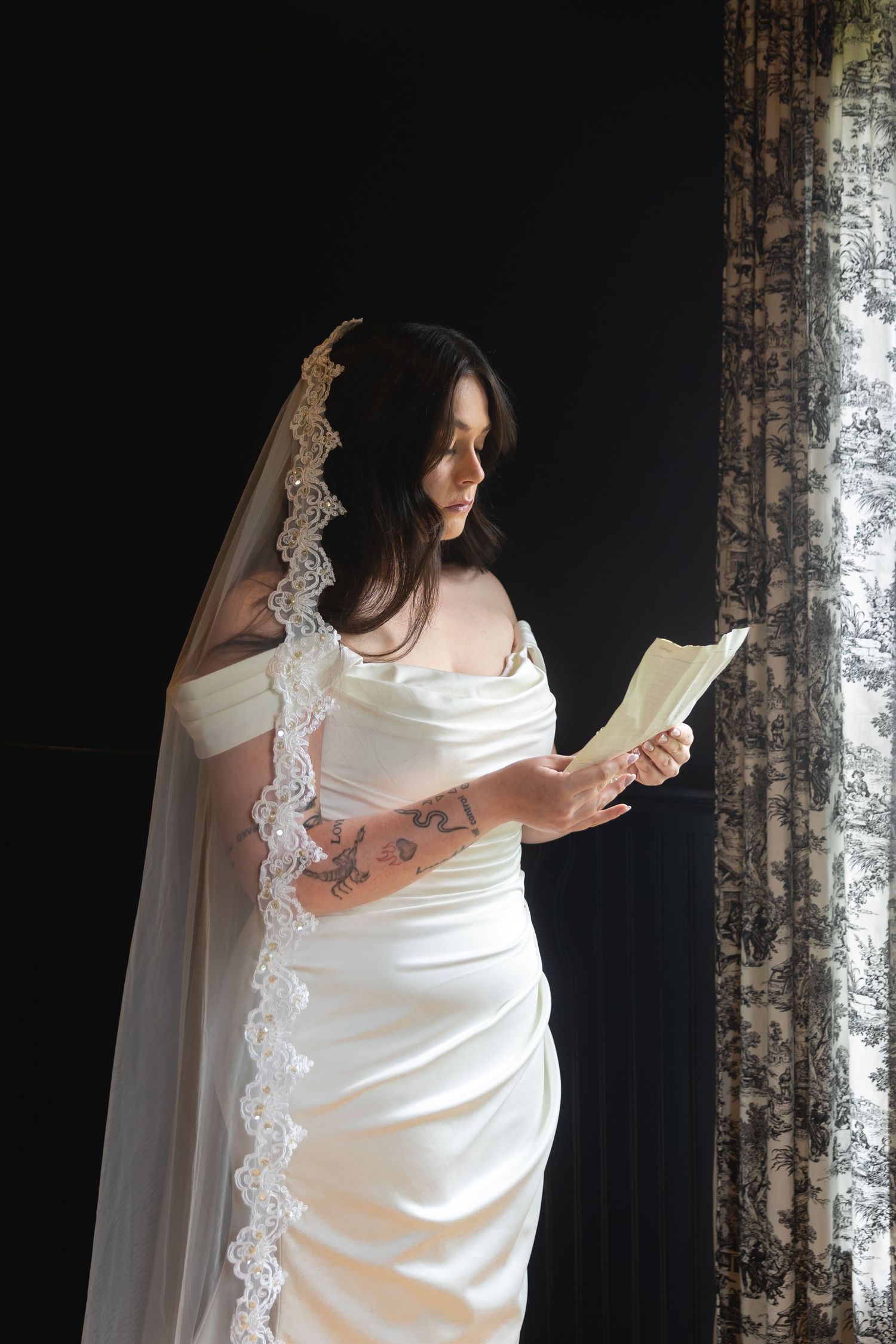 A bride in a white satin wedding dress with lace veil reads from a book near a window in dramatic lighting.