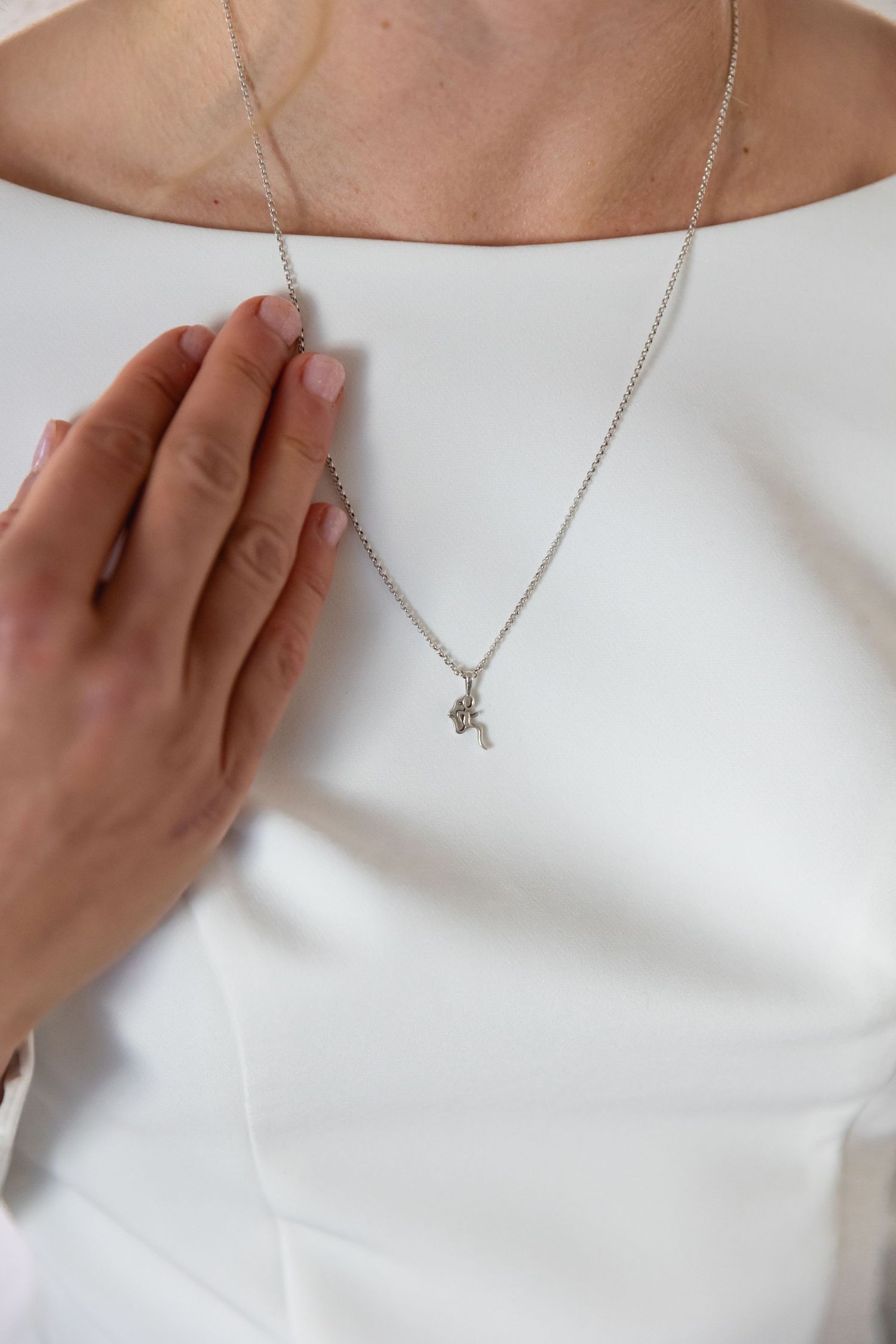 Close up of a delicate silver chain necklace with a small pendant being touched by a hand against a white top.