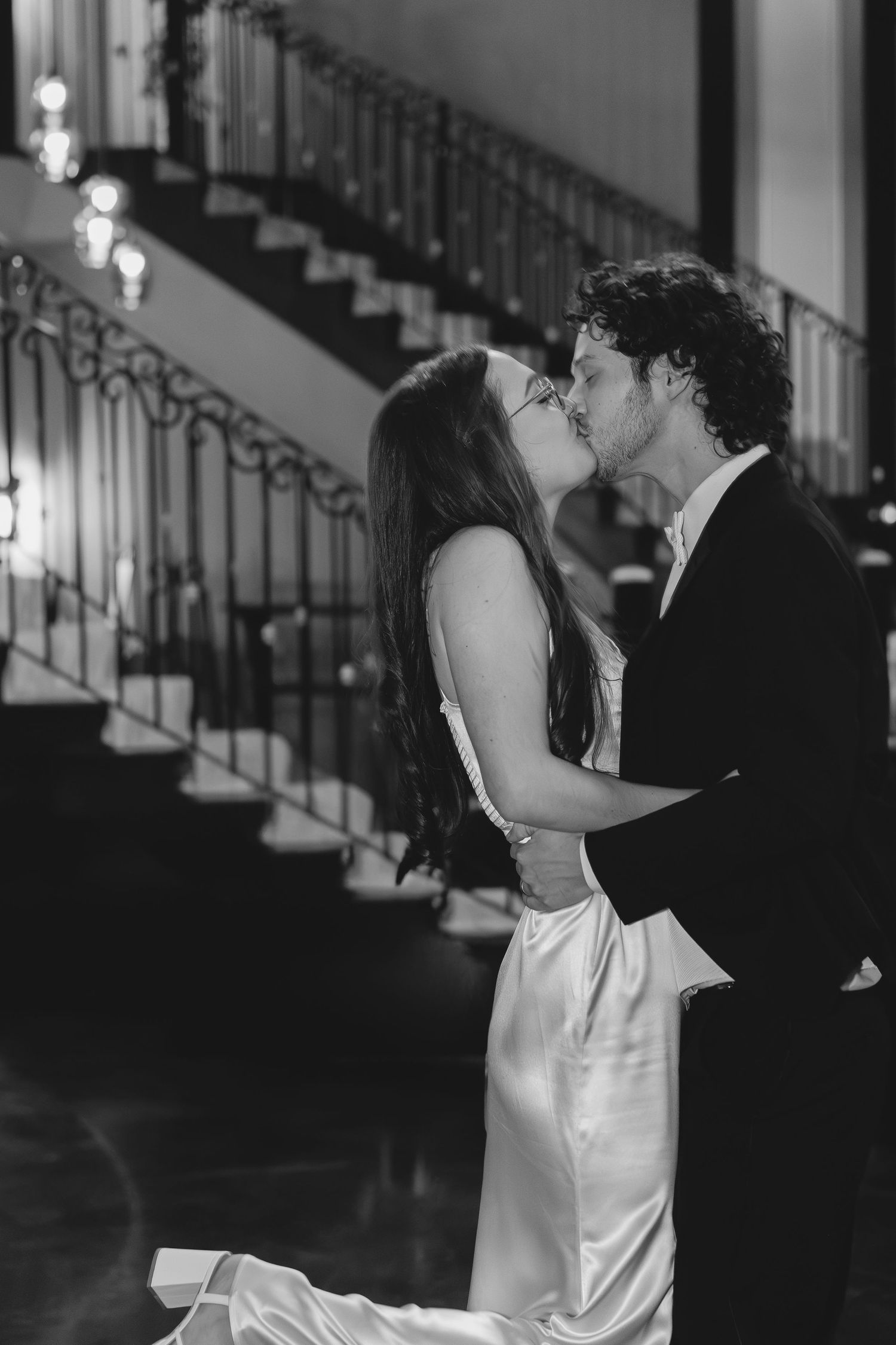 A romantic black and white moment with a couple embracing and kissing near an elegant staircase.