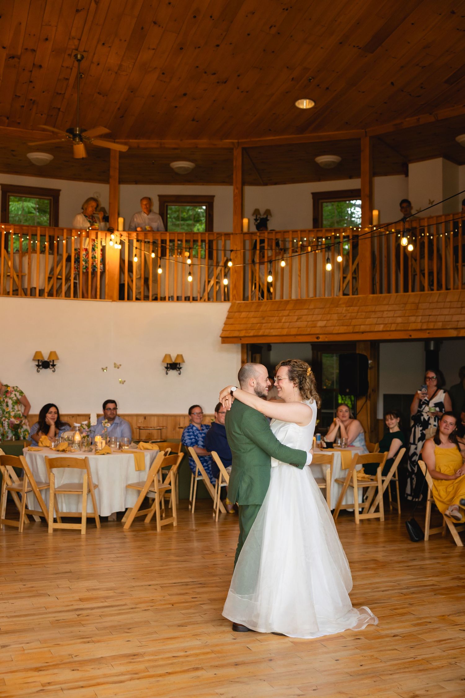 Couples dancing together in a rustic reception hall with wooden floors and balcony during a wedding celebration.