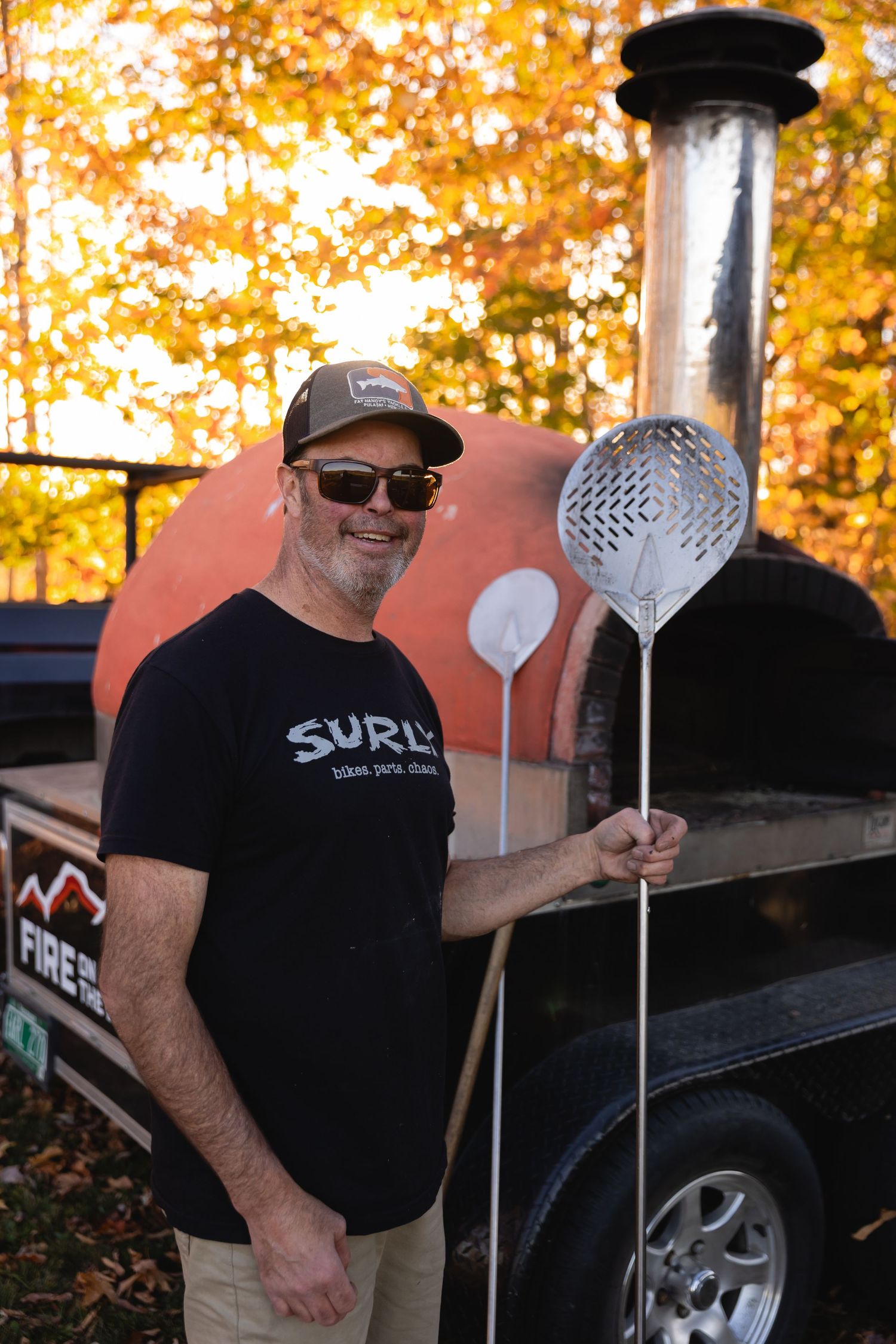 A person in a black Surly t-shirt and cap stands next to a grill holding cooking utensils on an autumn day.