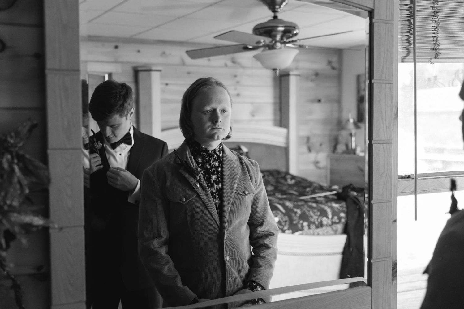 A black and white photograph shows two people standing in a rustic interior with a ceiling fan visible above.