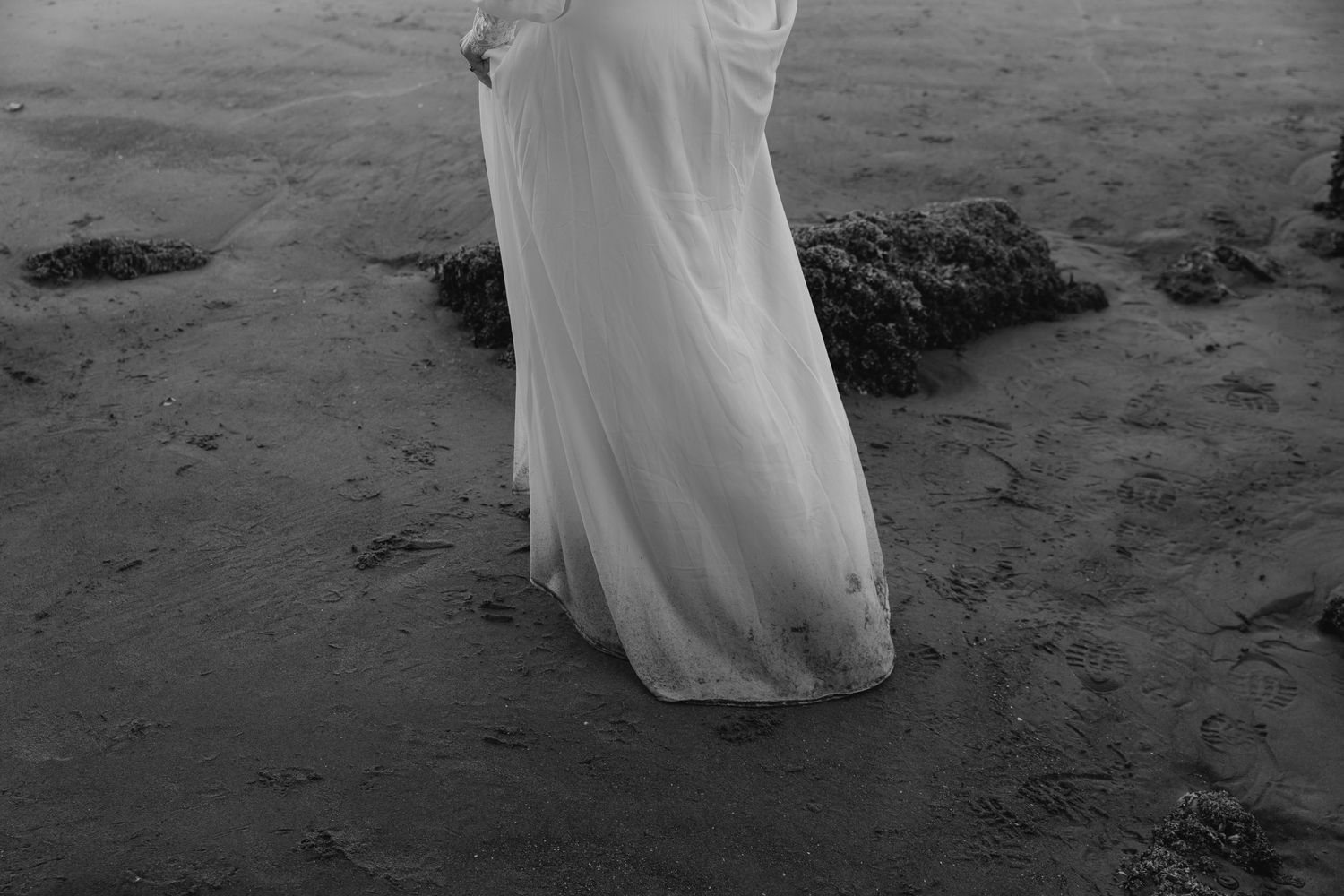 A bride in a flowing white dress walks along a rocky beach shoreline in black and white.