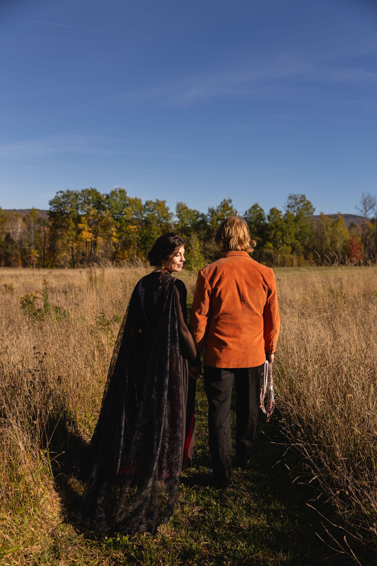 A couple walks through a golden autumn meadow with tall grass and trees in the background during sunset.