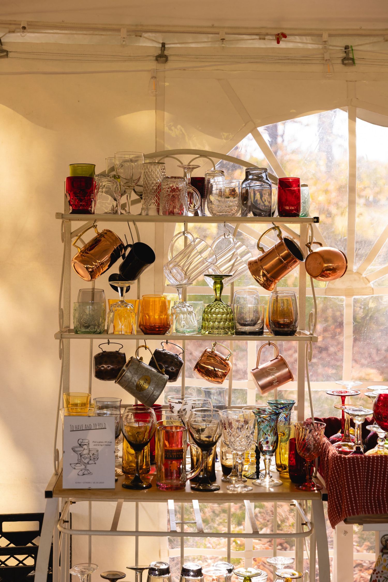 Vintage glassware and copper mugs displayed on illuminated glass shelves in a cozy sunlit corner.