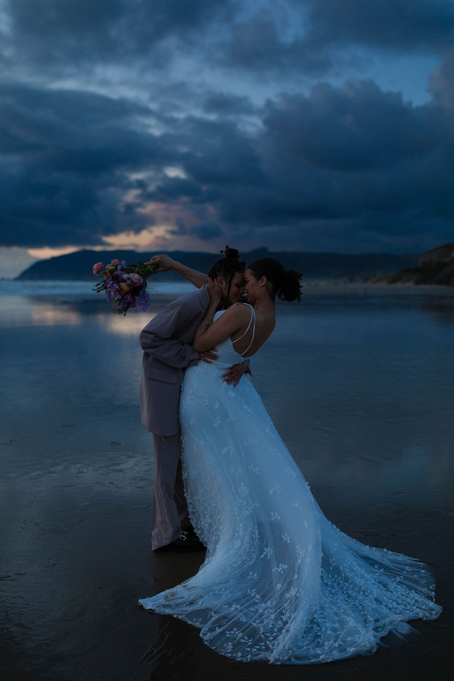 A couple shares a romantic moment on a beach with dramatic clouds and ocean reflections.