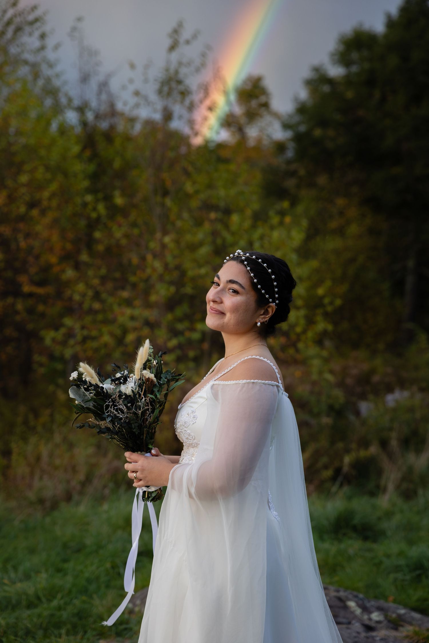 A romantic wedding portrait with a beautiful rainbow in the background above a wooded area.