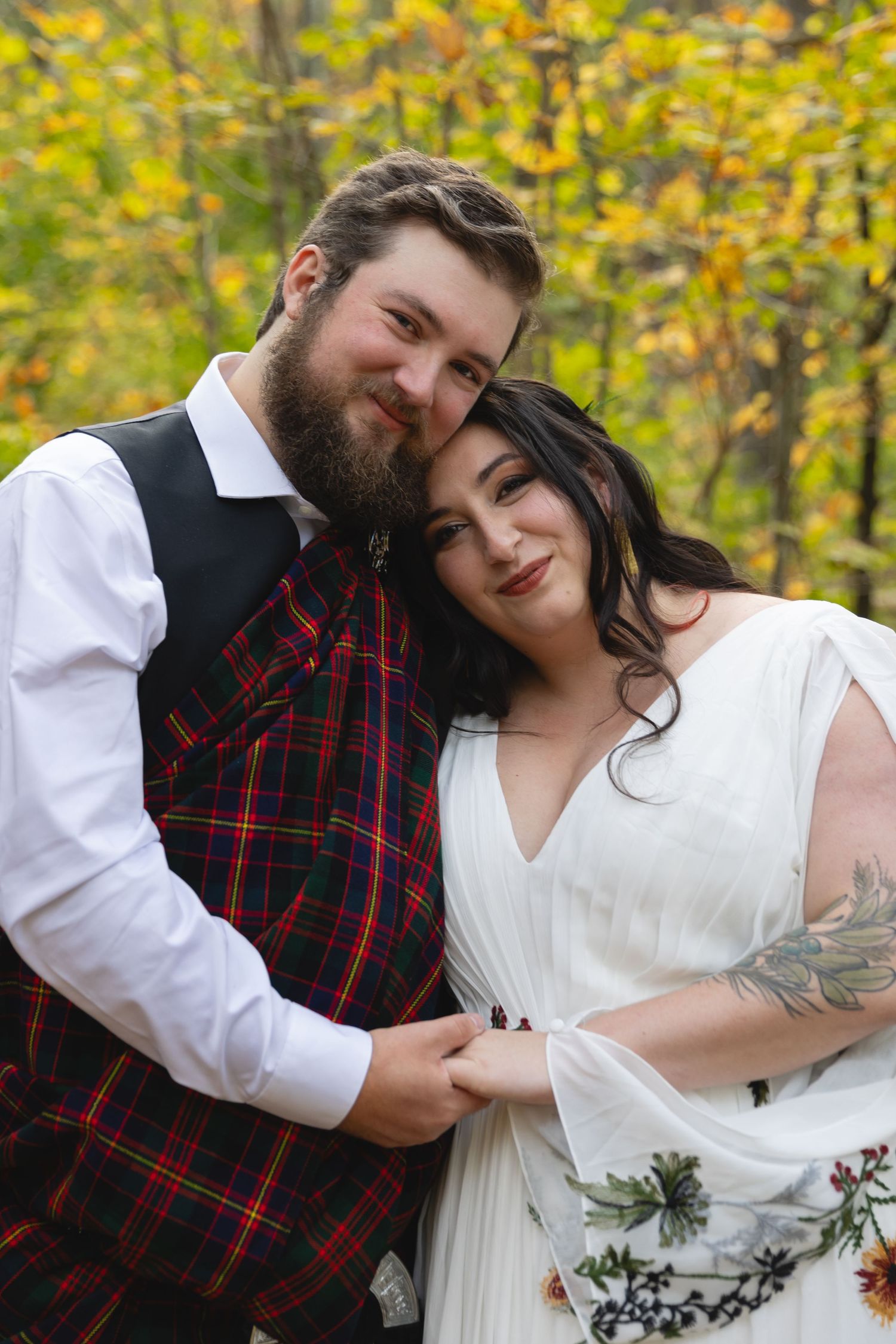 Happy couple in Scottish and traditional wedding attire pose against autumn forest backdrop.