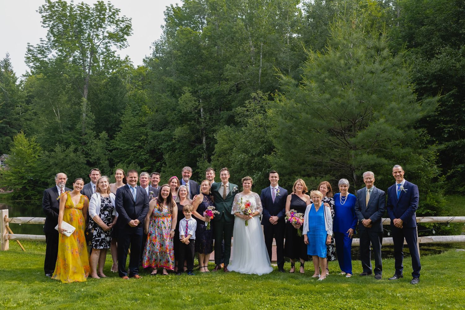 Large wedding party poses together outdoors with trees and greenery in the background.