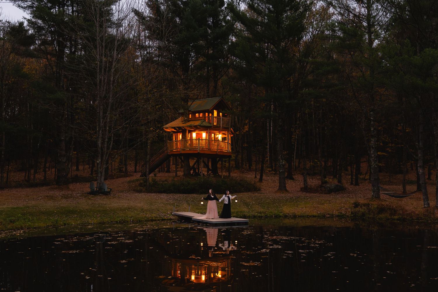 Wedding couple stands on dock reflected in pond before glowing treehouse at dusk.