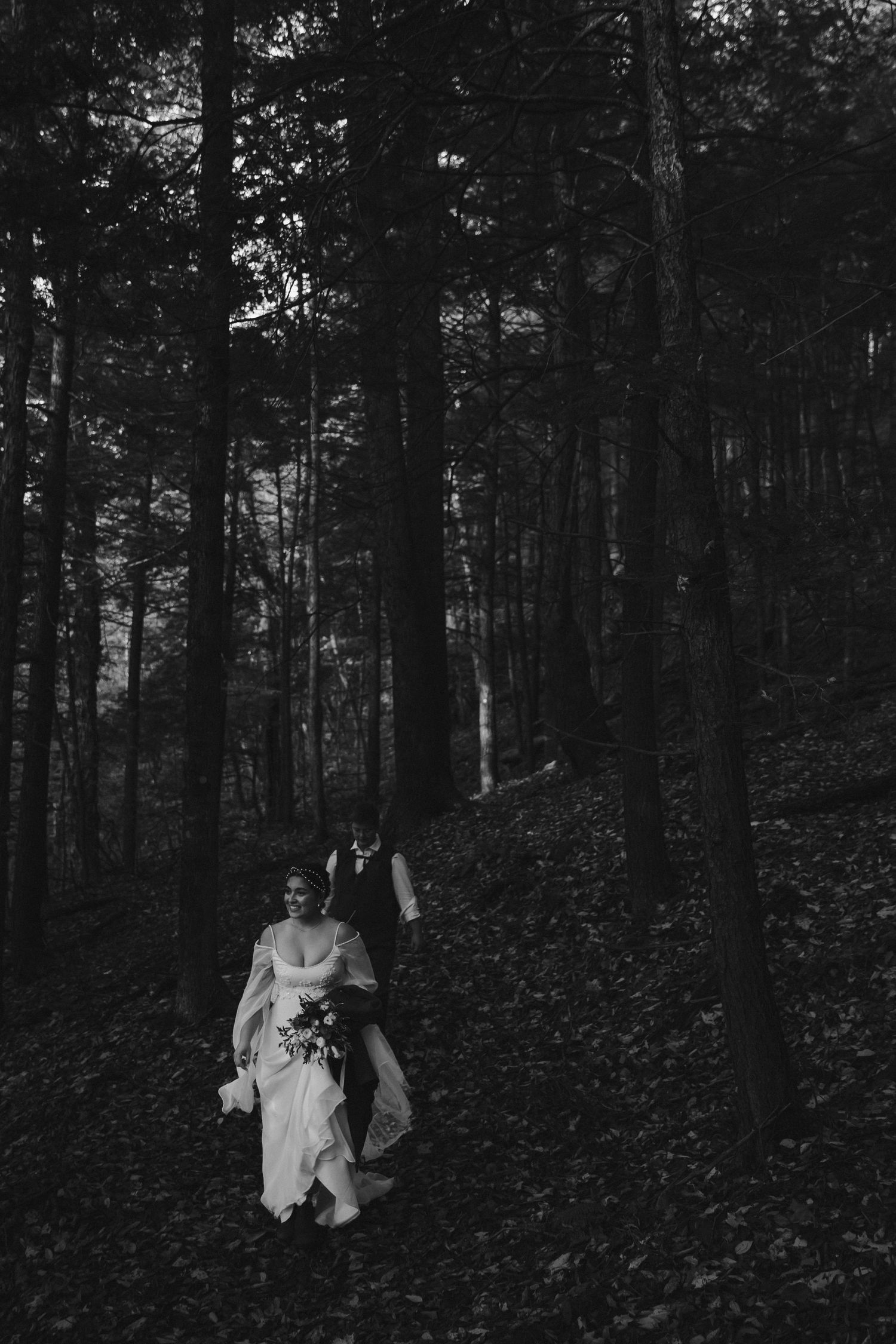 A couple walks down a forested path in formal attire surrounded by tall dark trees.