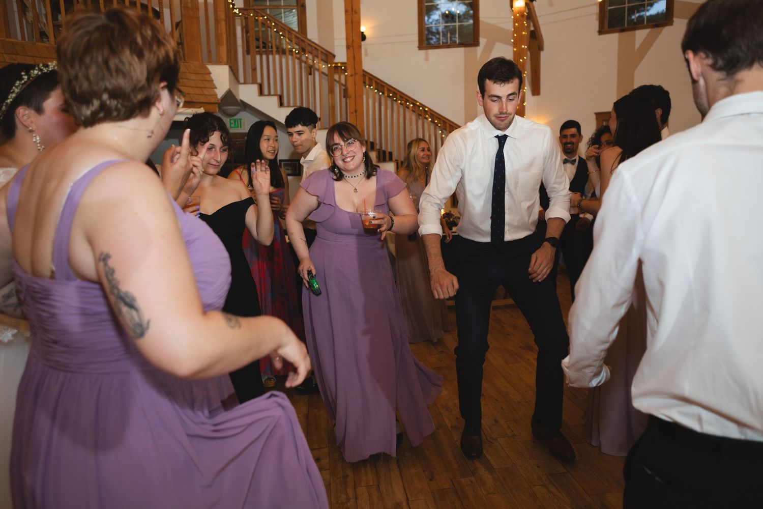 Reception guests in purple dresses and formal wear dancing together on a wooden floor.