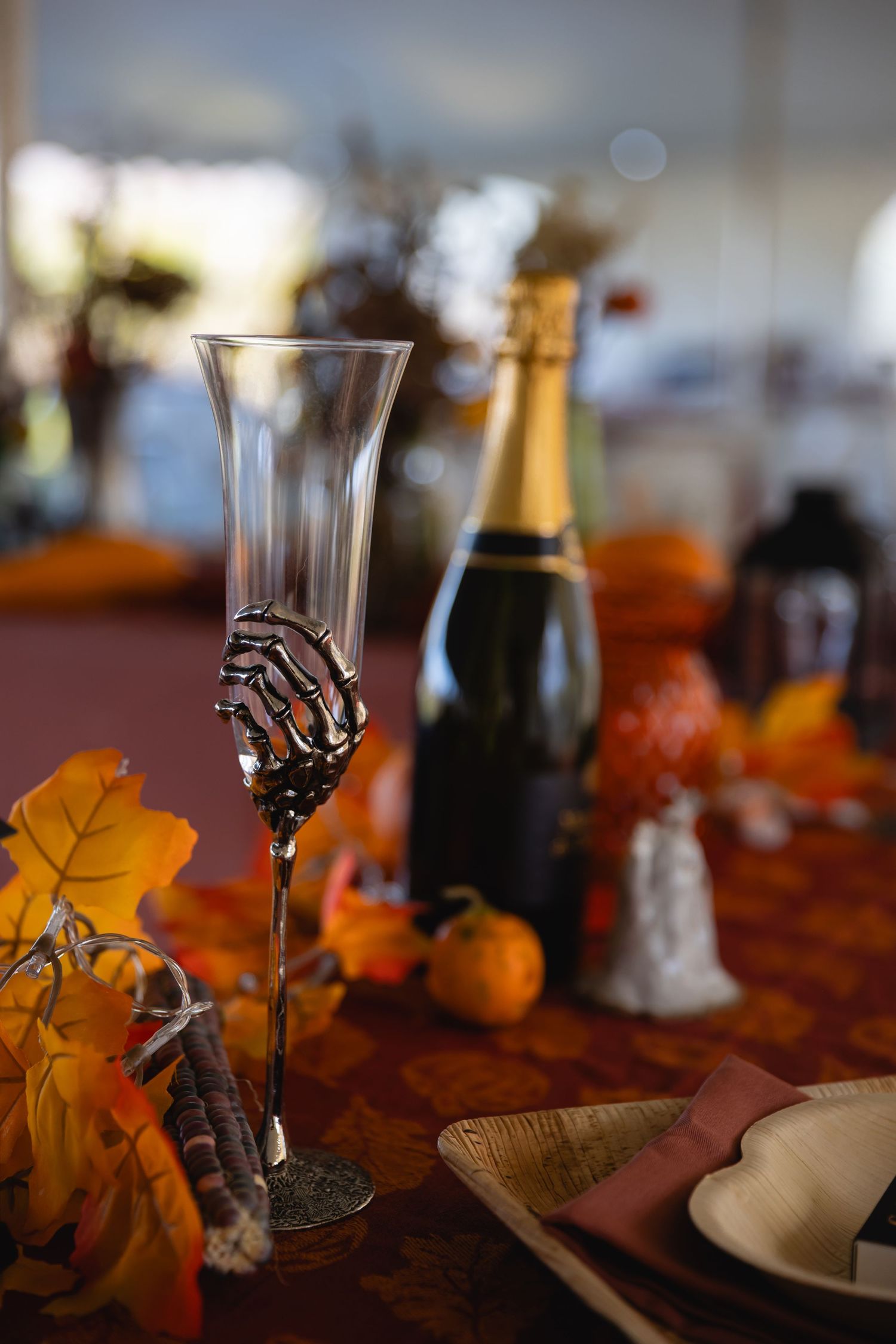 A Halloween-themed table setting with a skeleton hand champagne flute, orange leaves, and a bottle of bubbly.