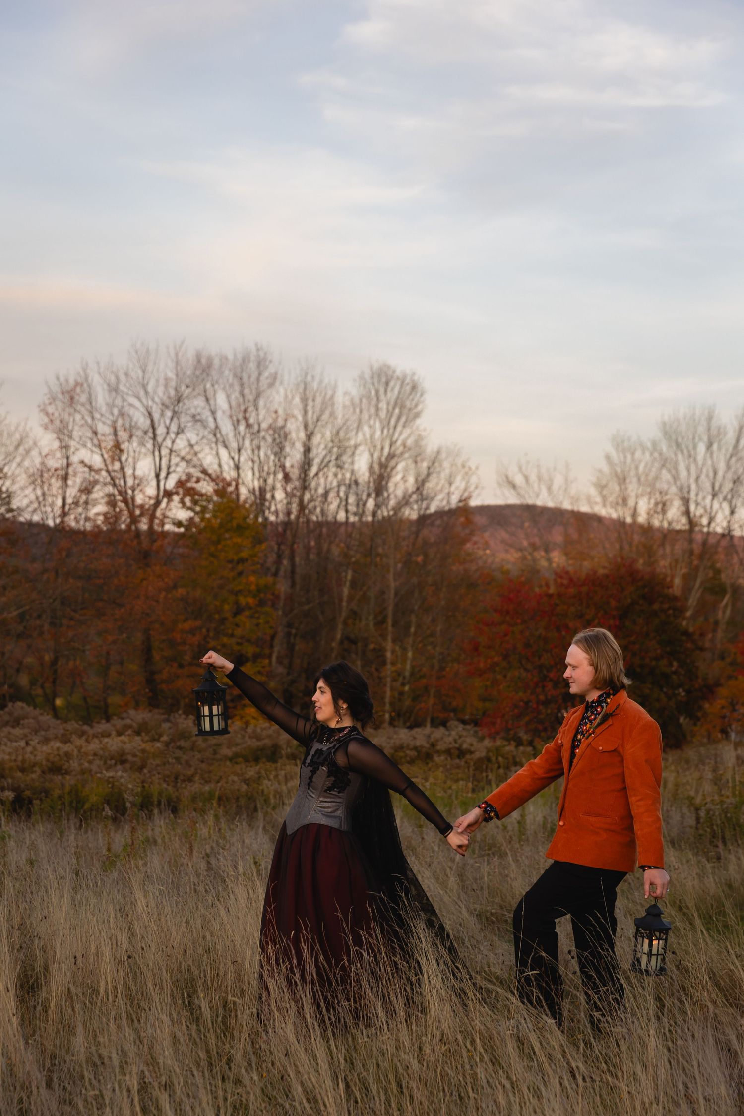 Two people hold hands while walking through a tall grass field at sunset during autumn with bare trees in background.