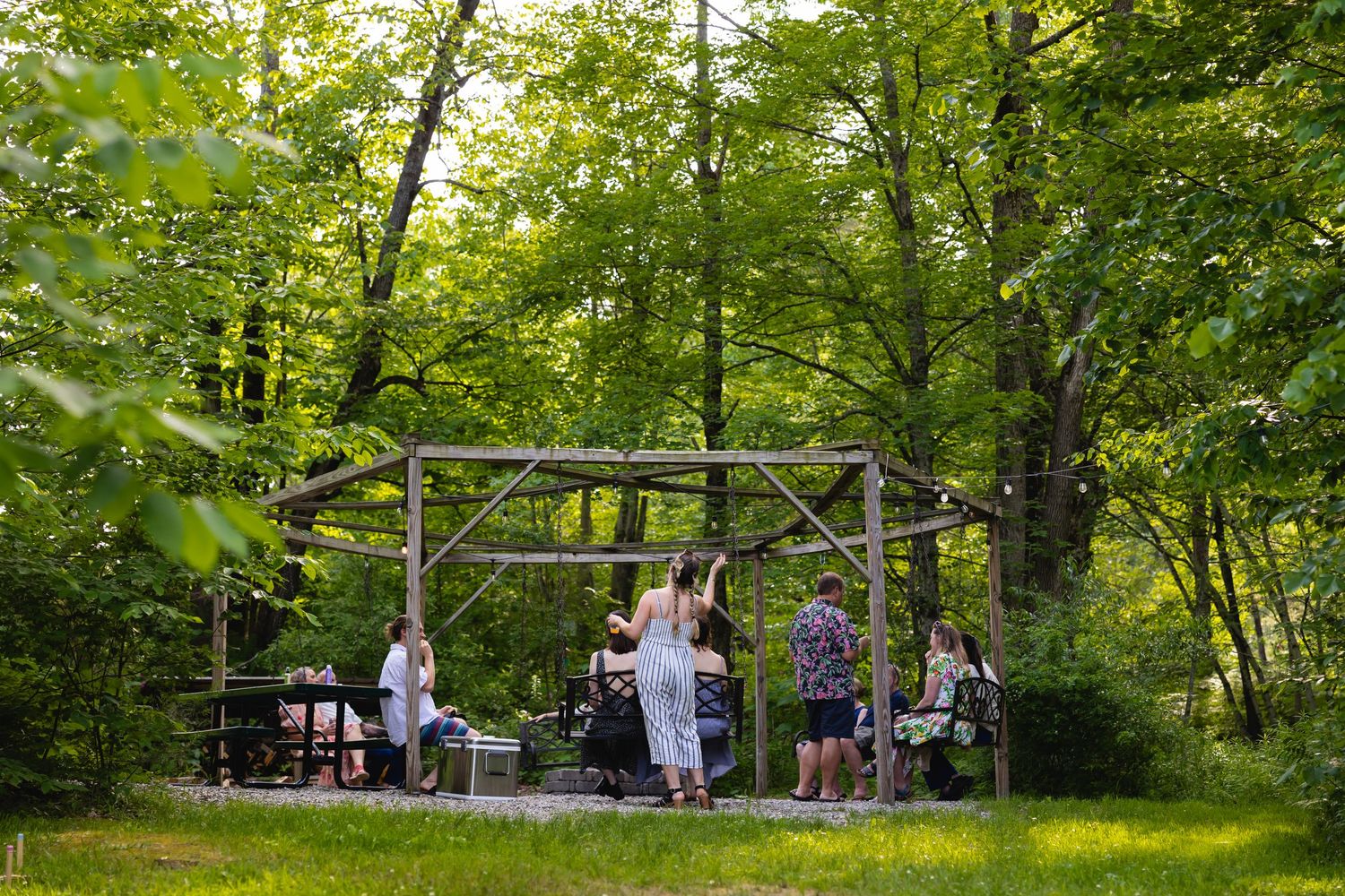 A group of people enjoy a casual outdoor gathering in a lush green forest setting with picnic tables and seating.