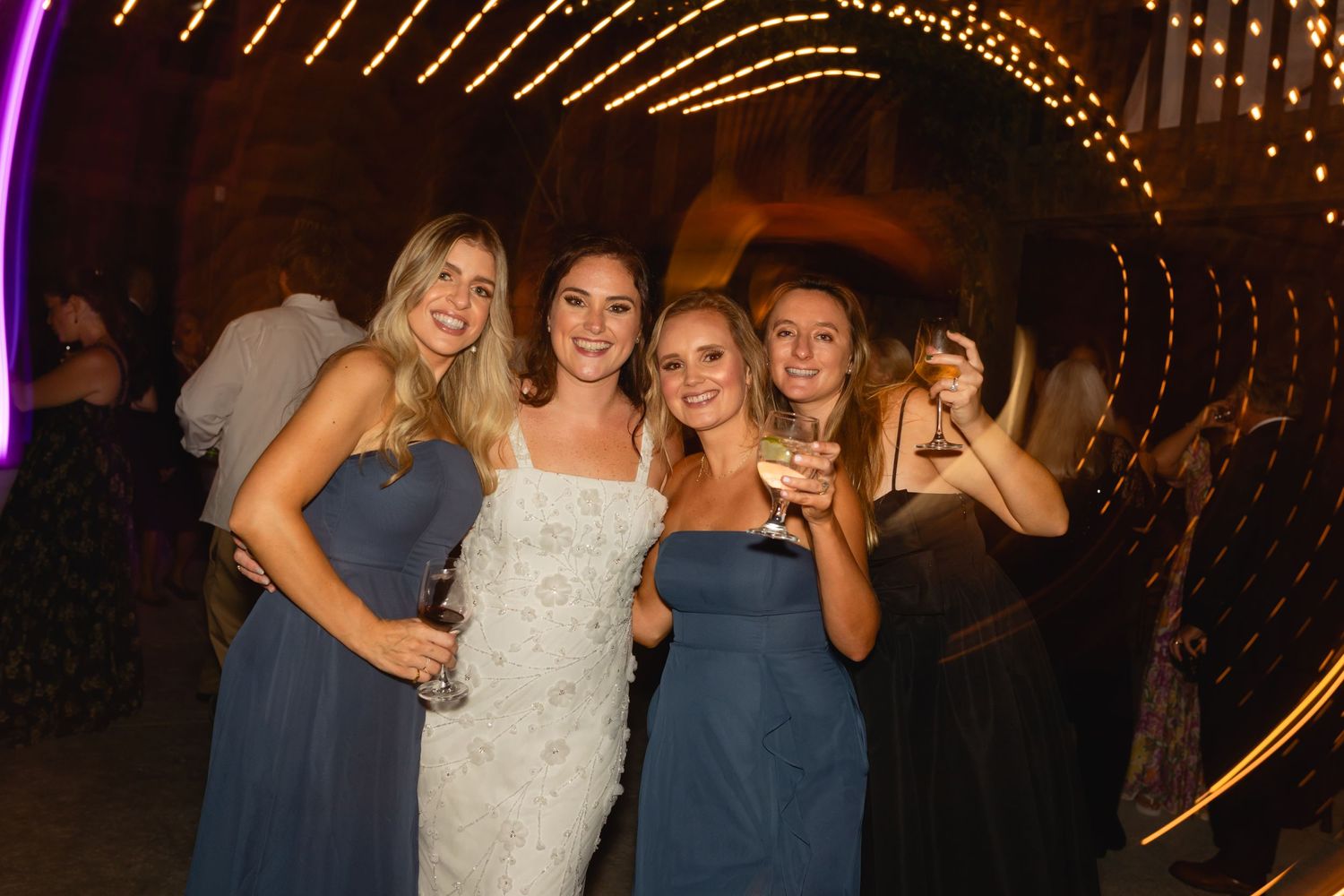Wedding celebration in a tunnel decorated with string lights as group poses together in blue dresses.