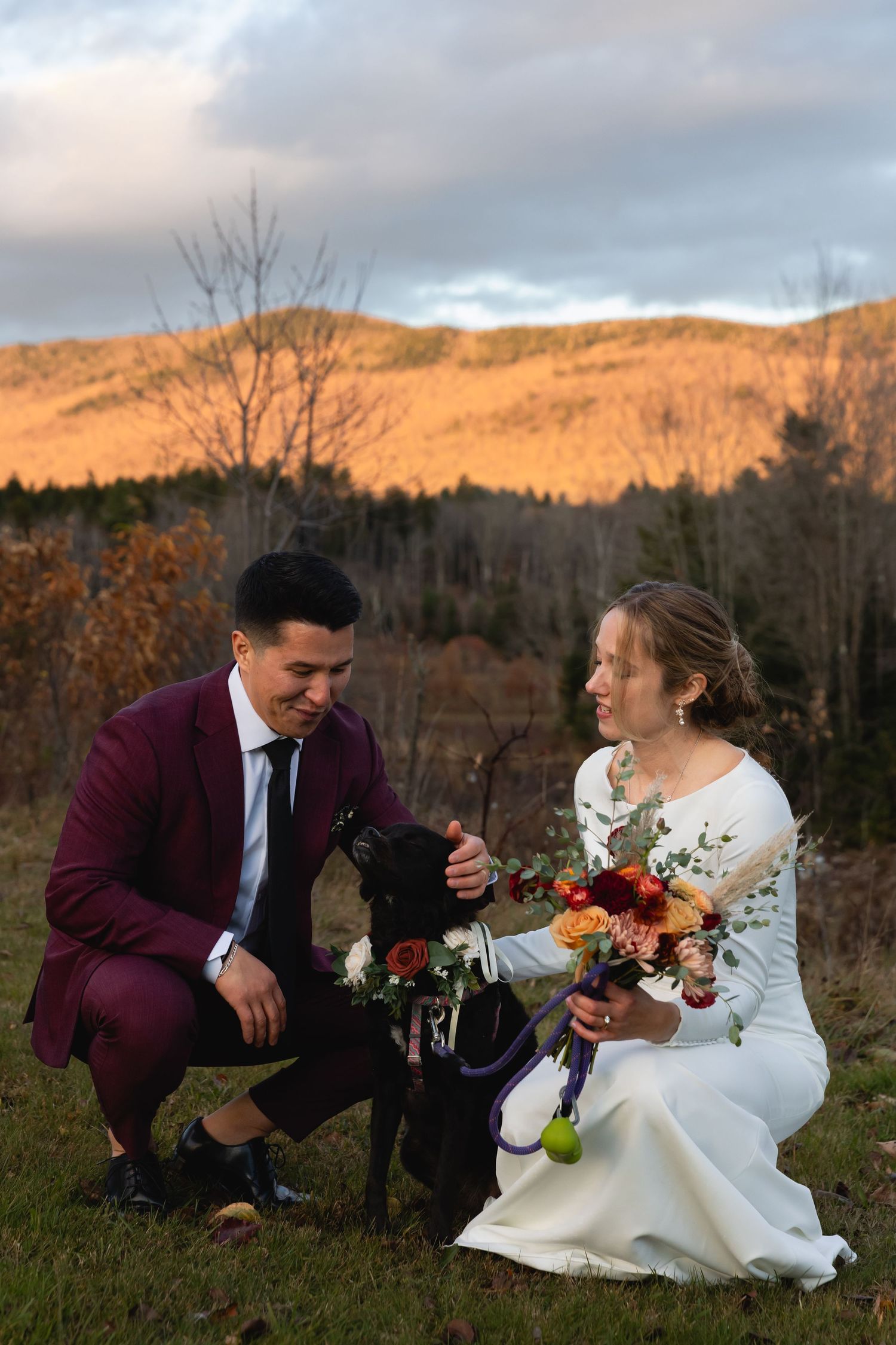 A couple in wedding attire kneels on grass at sunset with mountains and autumn trees in the background.