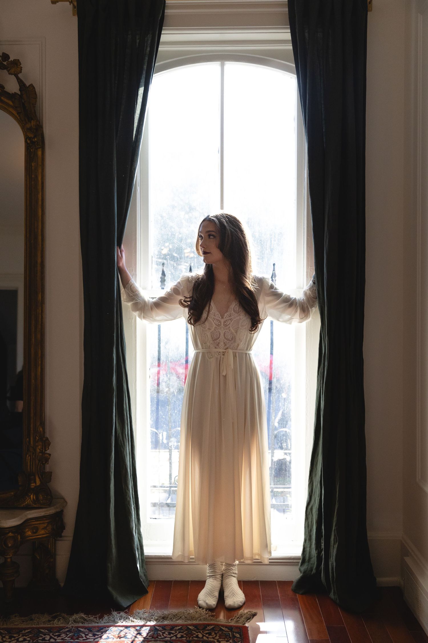 Figure in a flowing white vintage dress stands backlit at tall window with dark curtains and ornate mirror on wall.