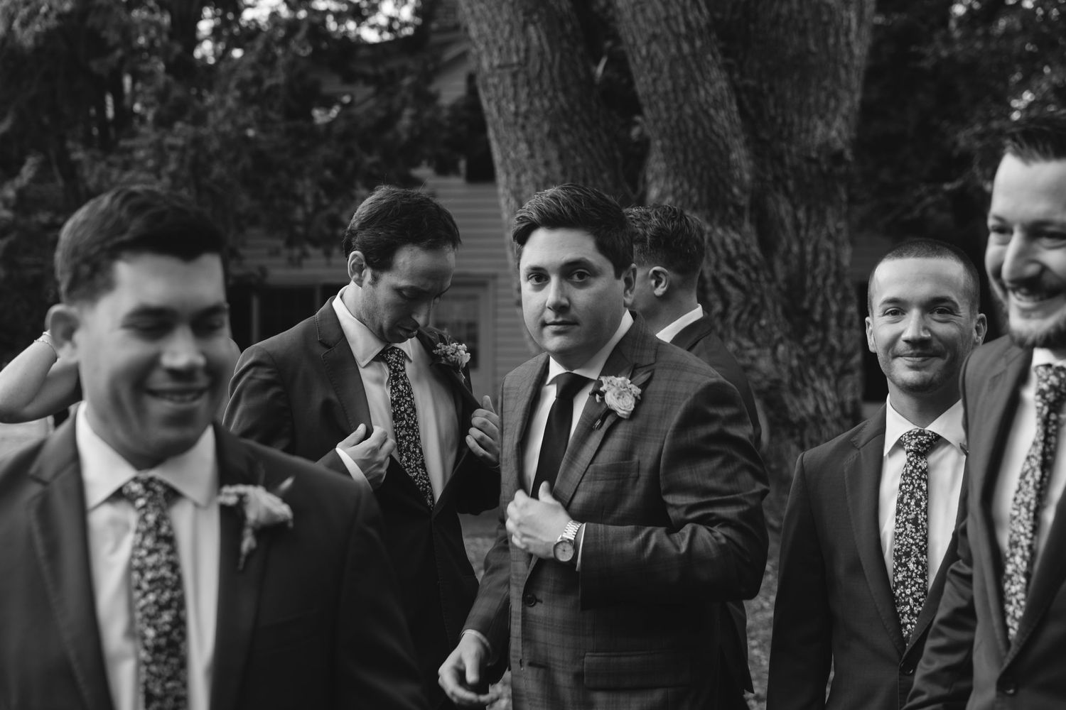 Black and white candid photograph of a wedding party in suits and ties laughing together outdoors.