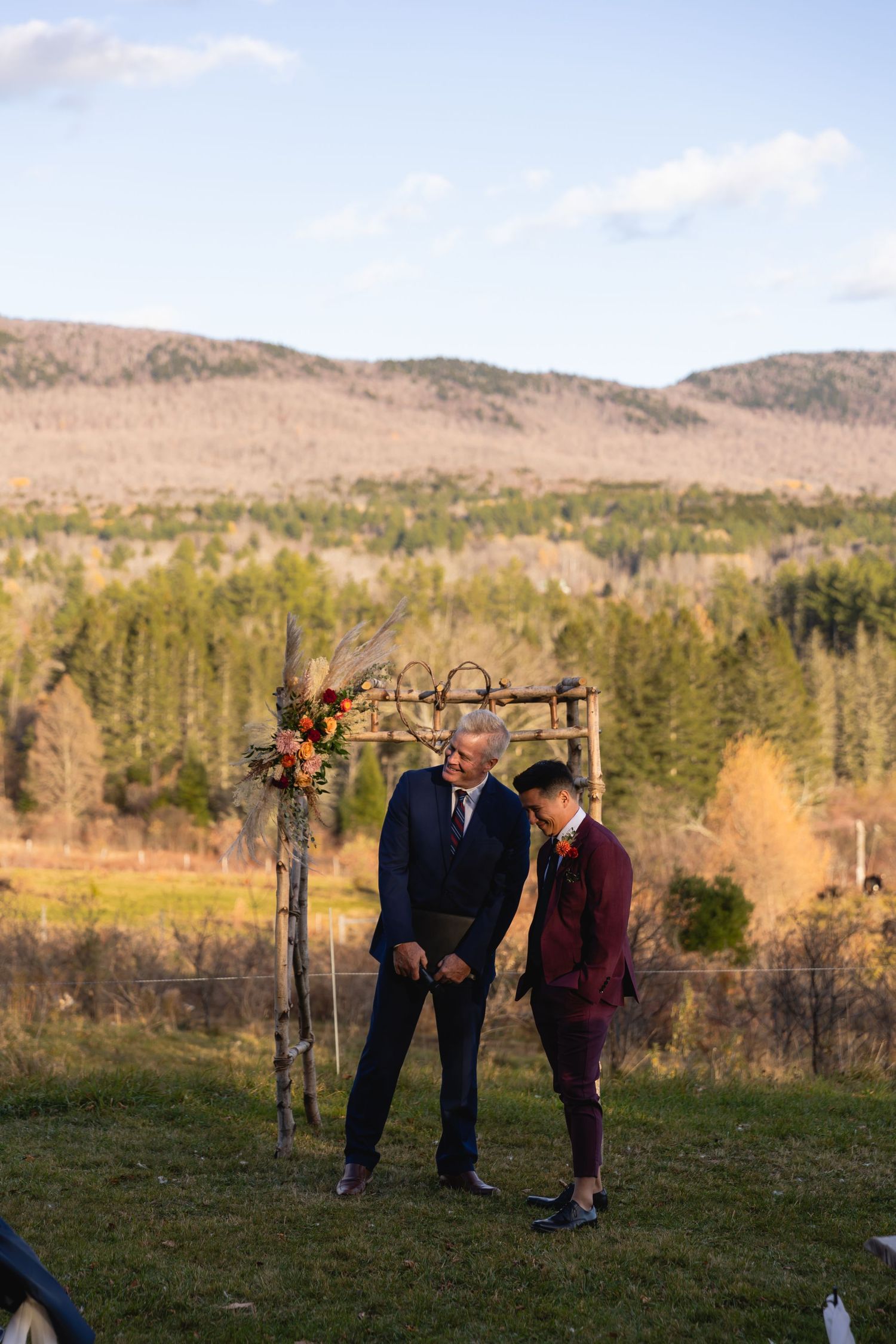 A couple stands together in an outdoor mountain setting at sunset beneath a rustic wooden arch in autumn.