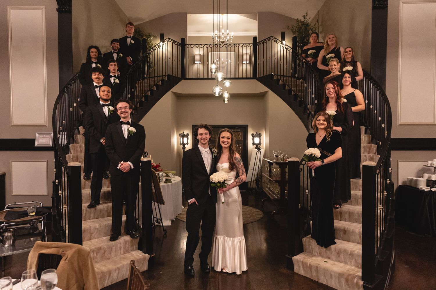 Wedding party posed on an elegant curved staircase in formal attire inside a classic venue with warm lighting.