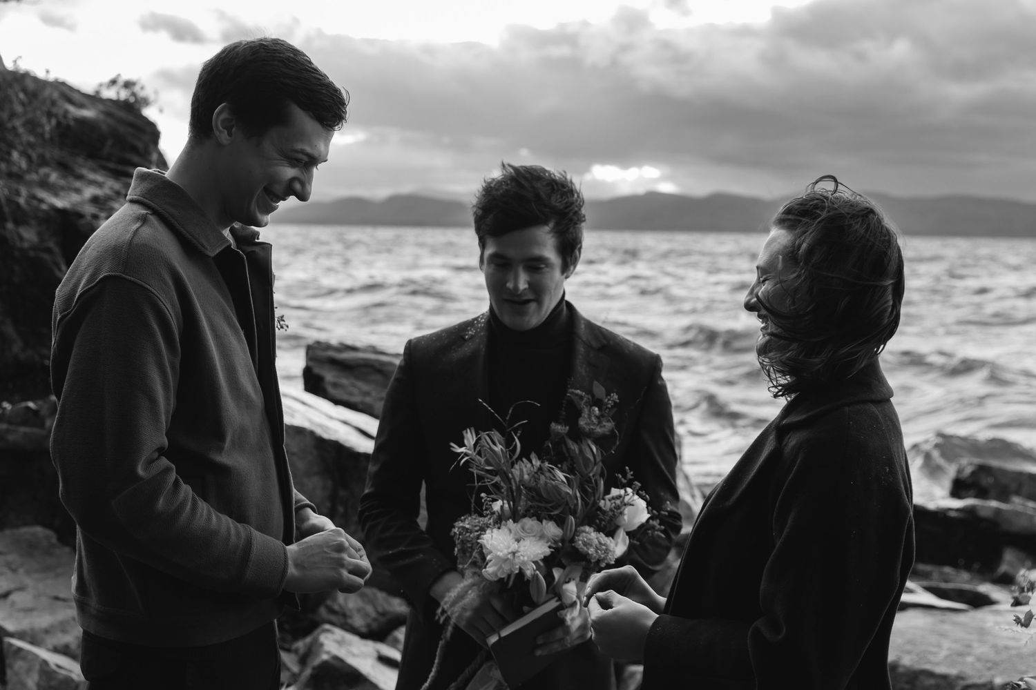 A vintage black and white coastal photo shows three people gathered near crashing waves on a rocky shoreline.