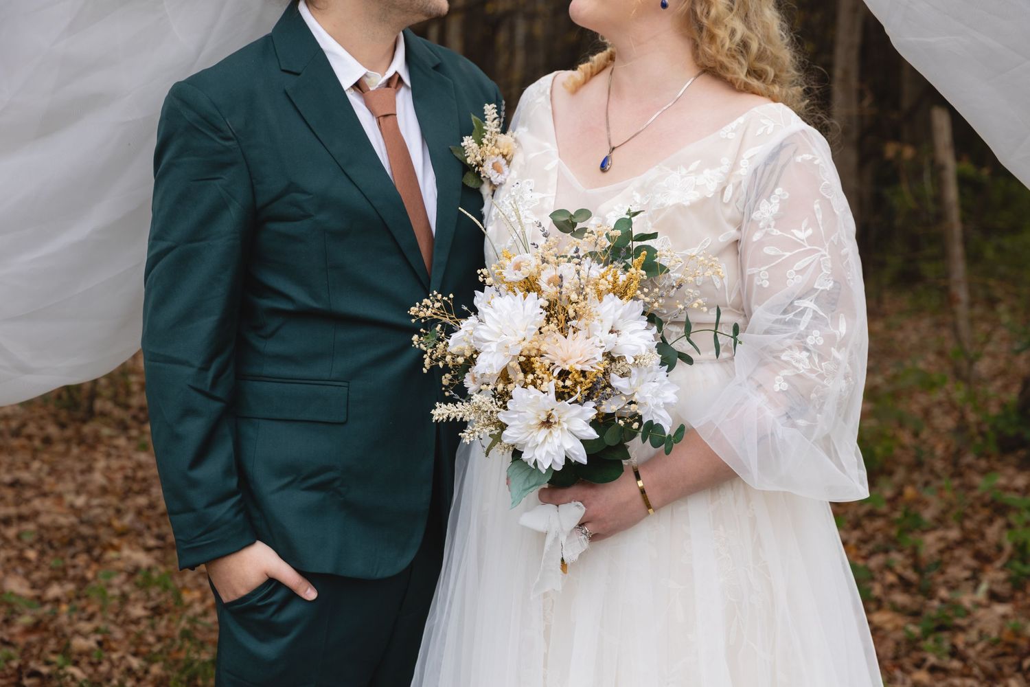 Elegant wedding attire featuring emerald suit and lace dress with white and yellow bouquet.