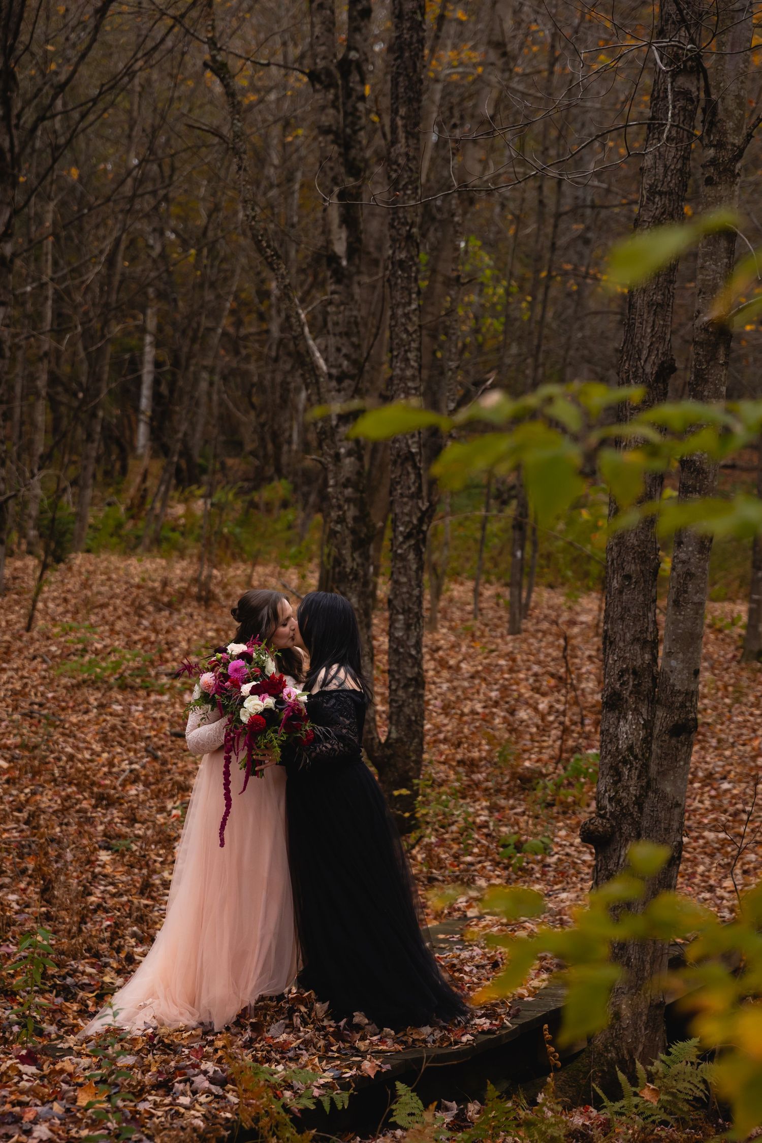 Two brides kissing in autumn forest, one in pink tulle gown and one in black dress with bouquet.