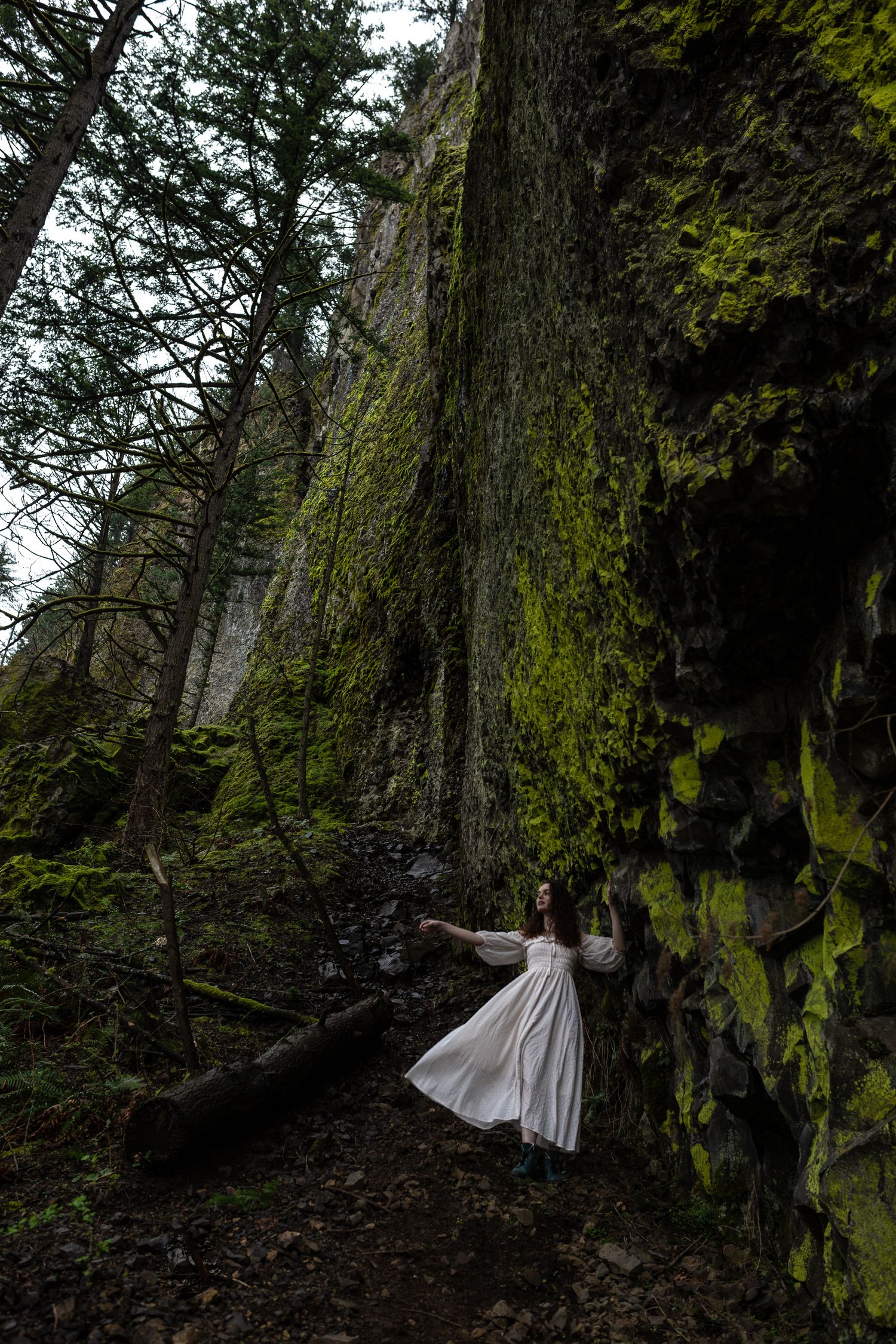 Woman in white flowing dress leaning against moss-covered rock cliff in dark forest.