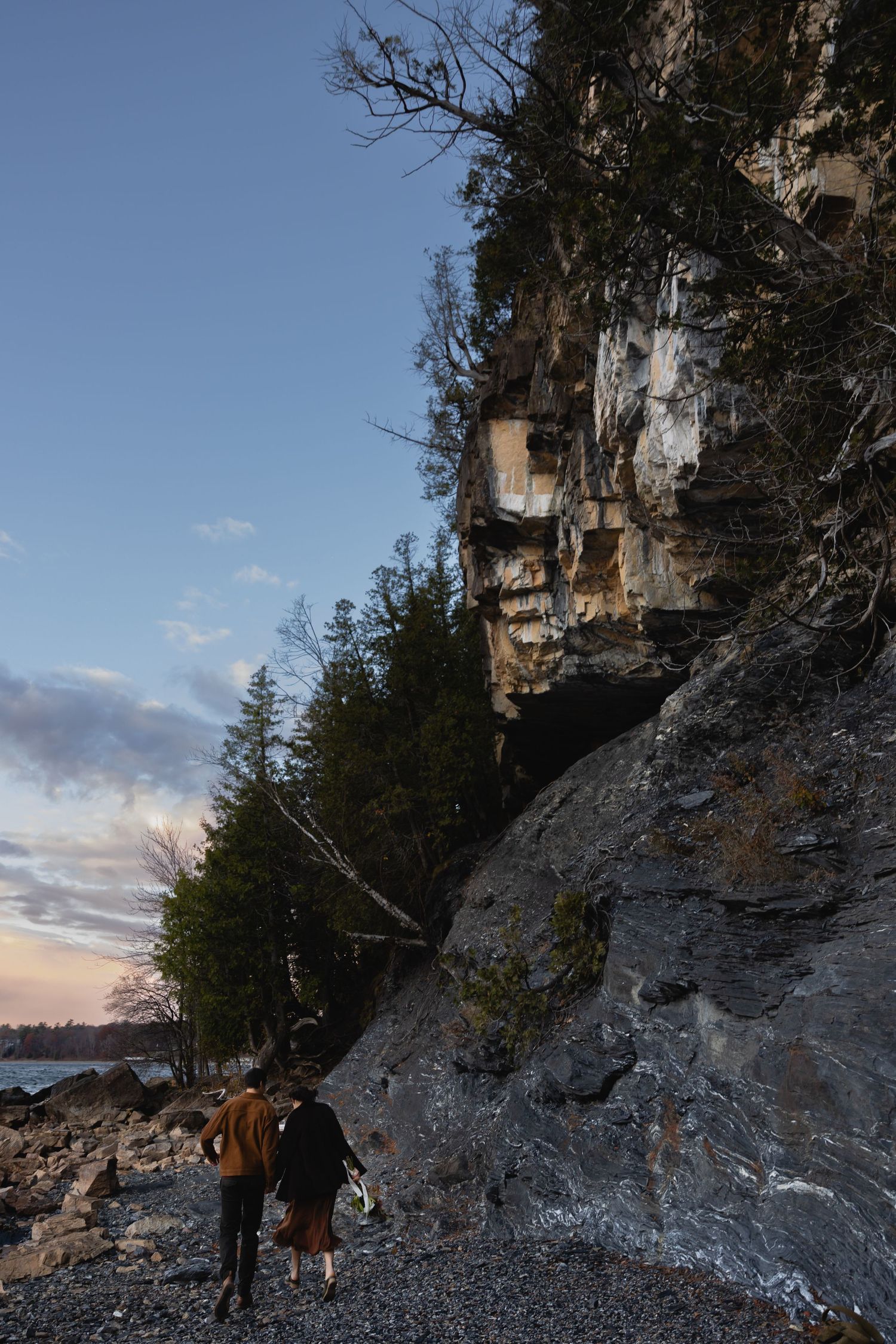 Two hikers walking along a rocky shoreline beneath towering limestone cliffs at sunset with evergreen trees overhead.