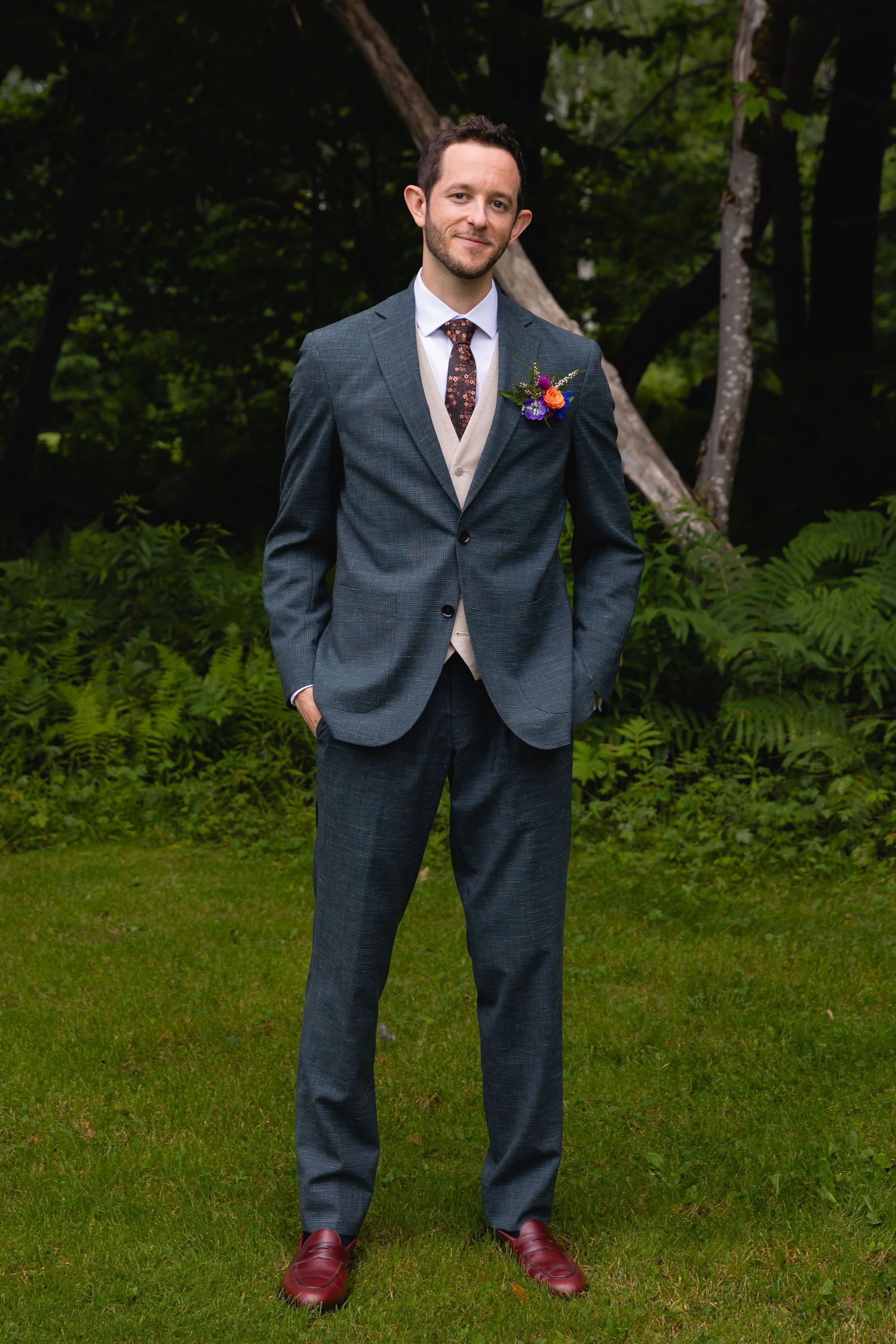 Groom in tailored gray suit with burgundy tie and boutonniere standing in garden setting.